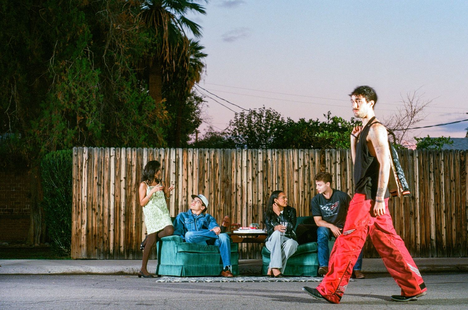 Group of young people gathered on urban street at dusk near wooden fence.