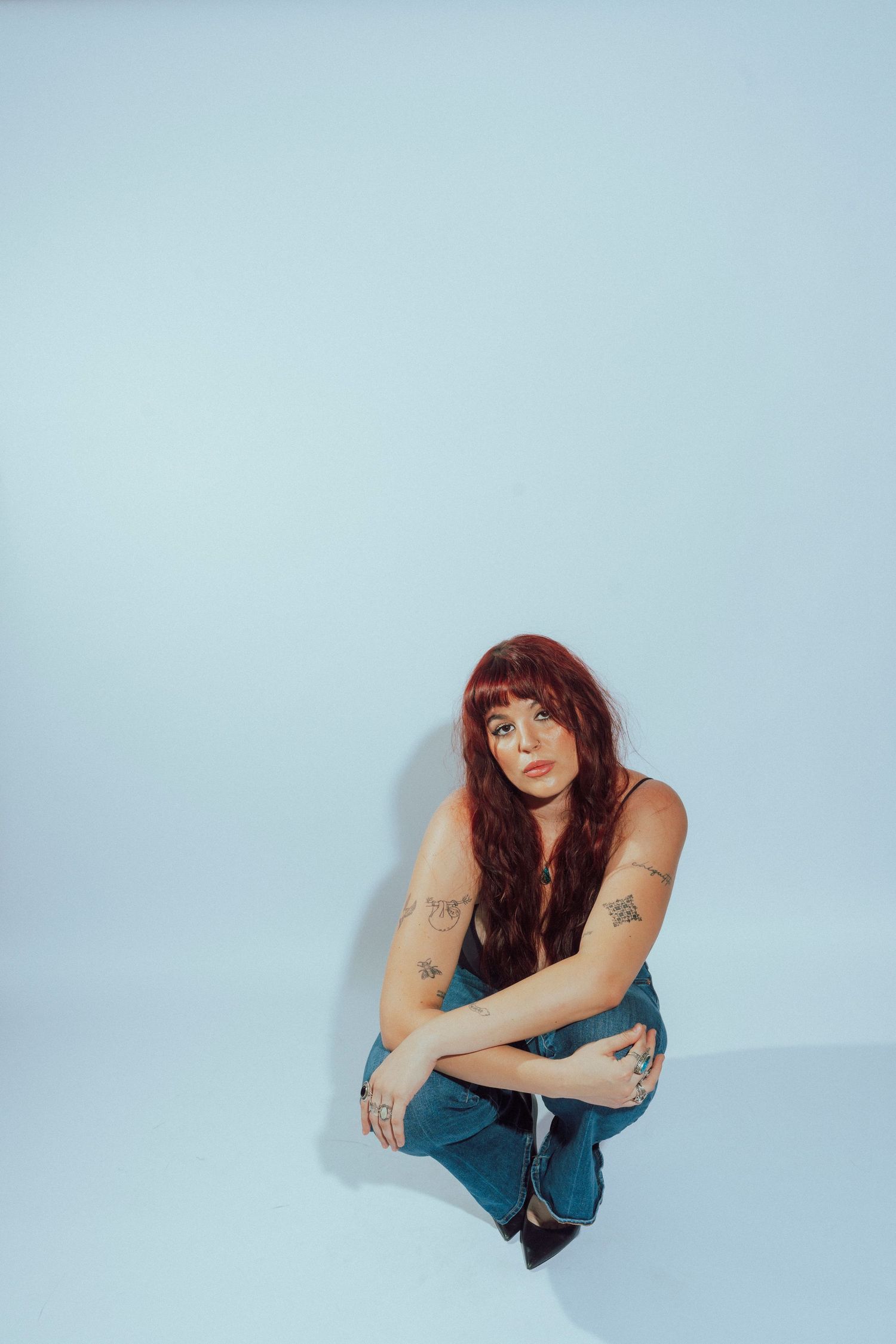 Young woman with red hair and tattoos sitting on floor in jeans against light blue background.
