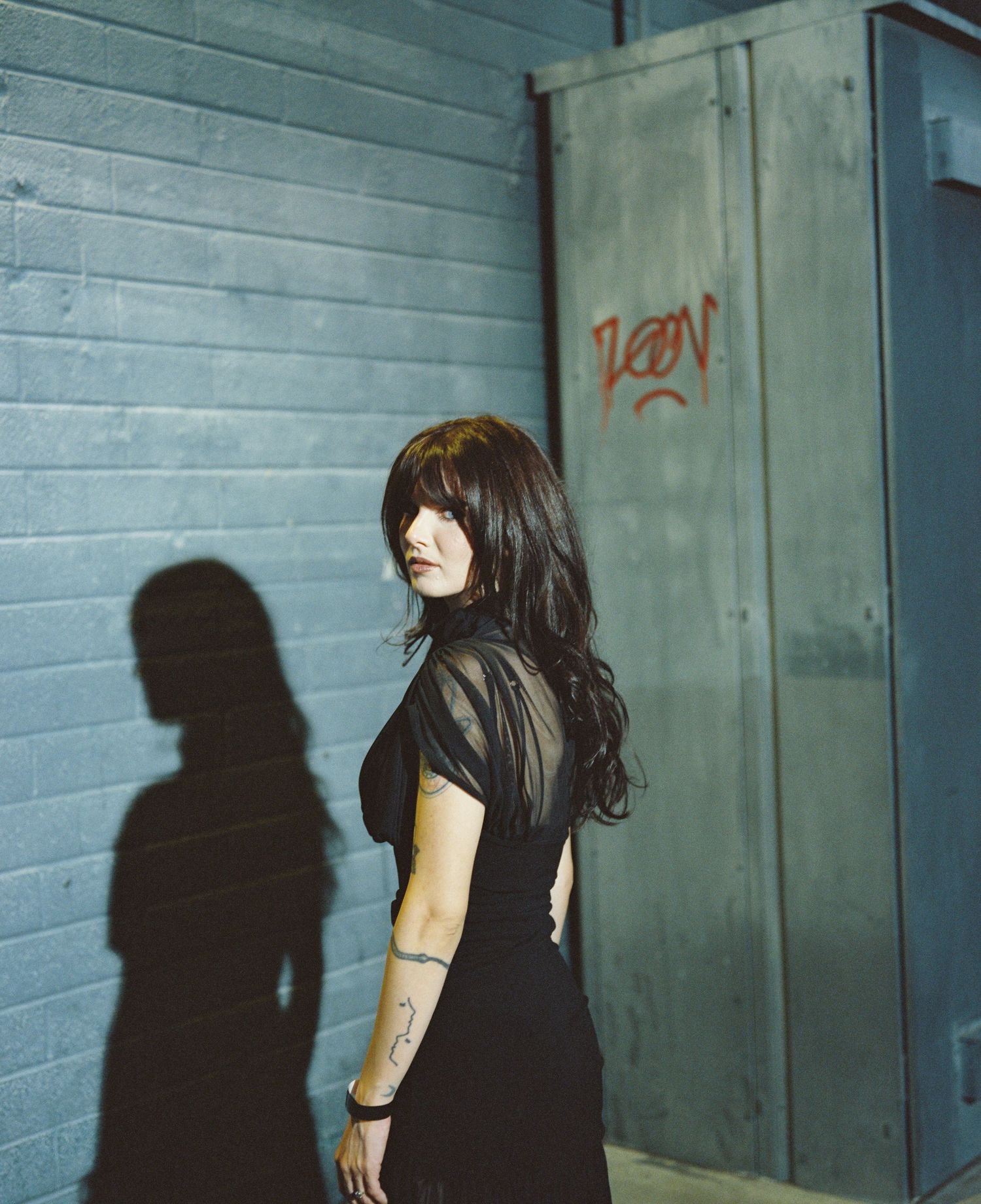 Woman with long dark hair in black outfit standing by gray wall with graffiti.