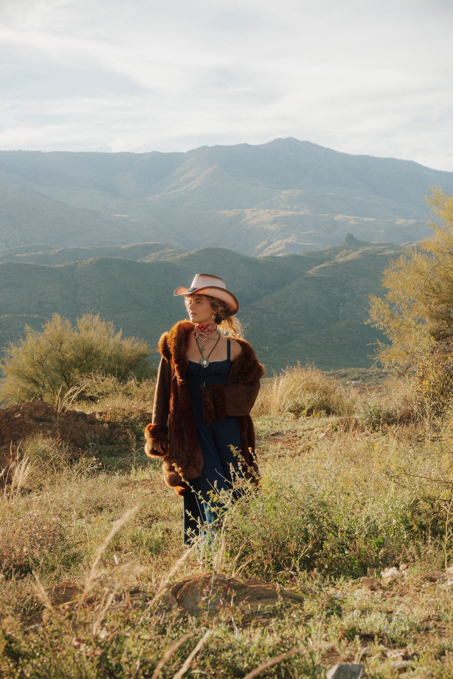 Woman in cowboy hat and fur coat stands on hillside overlooking scenic green mountain valley.