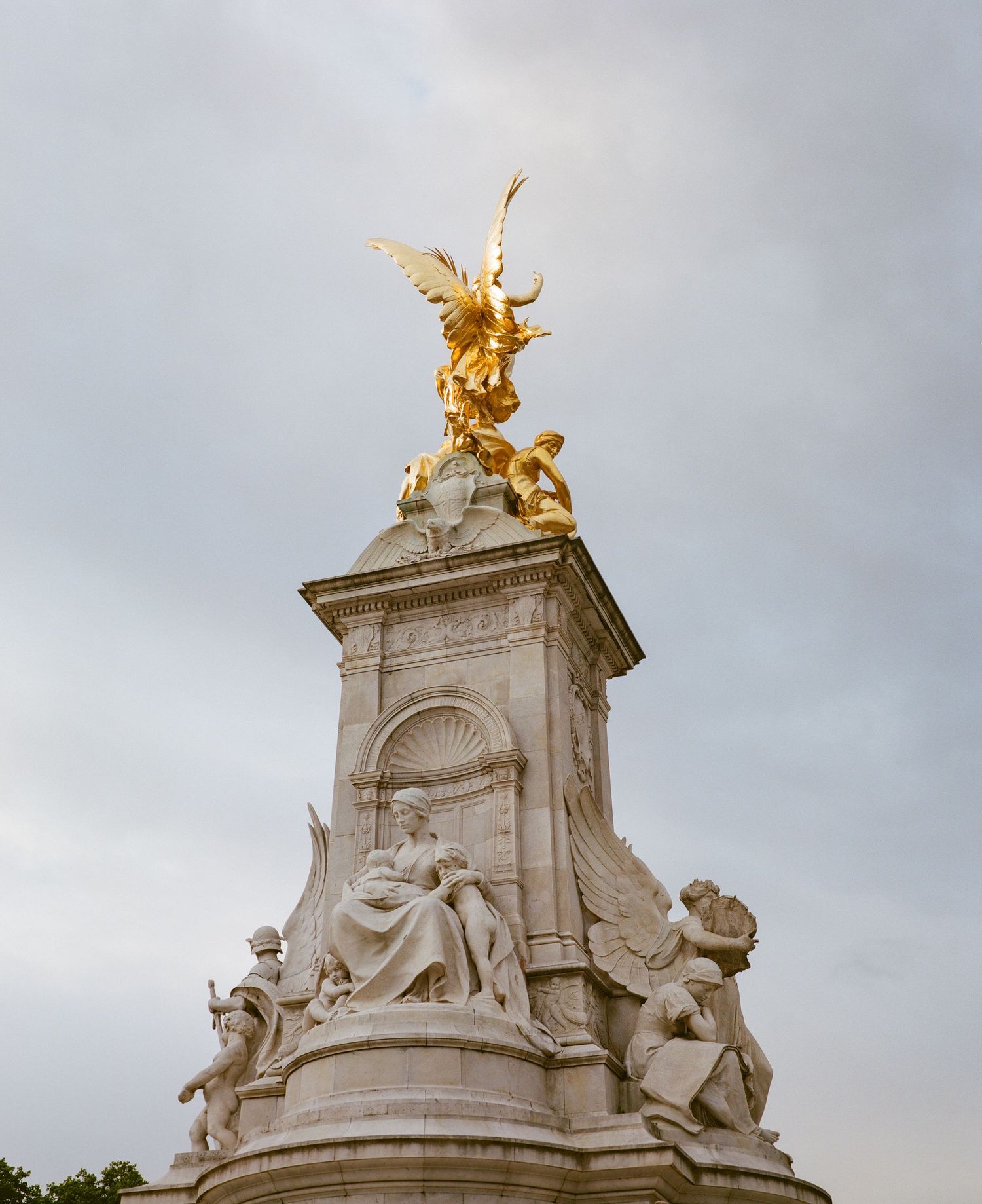 Golden winged statue atop the Victoria Memorial monument with white marble sculptures against a cloudy sky in London.