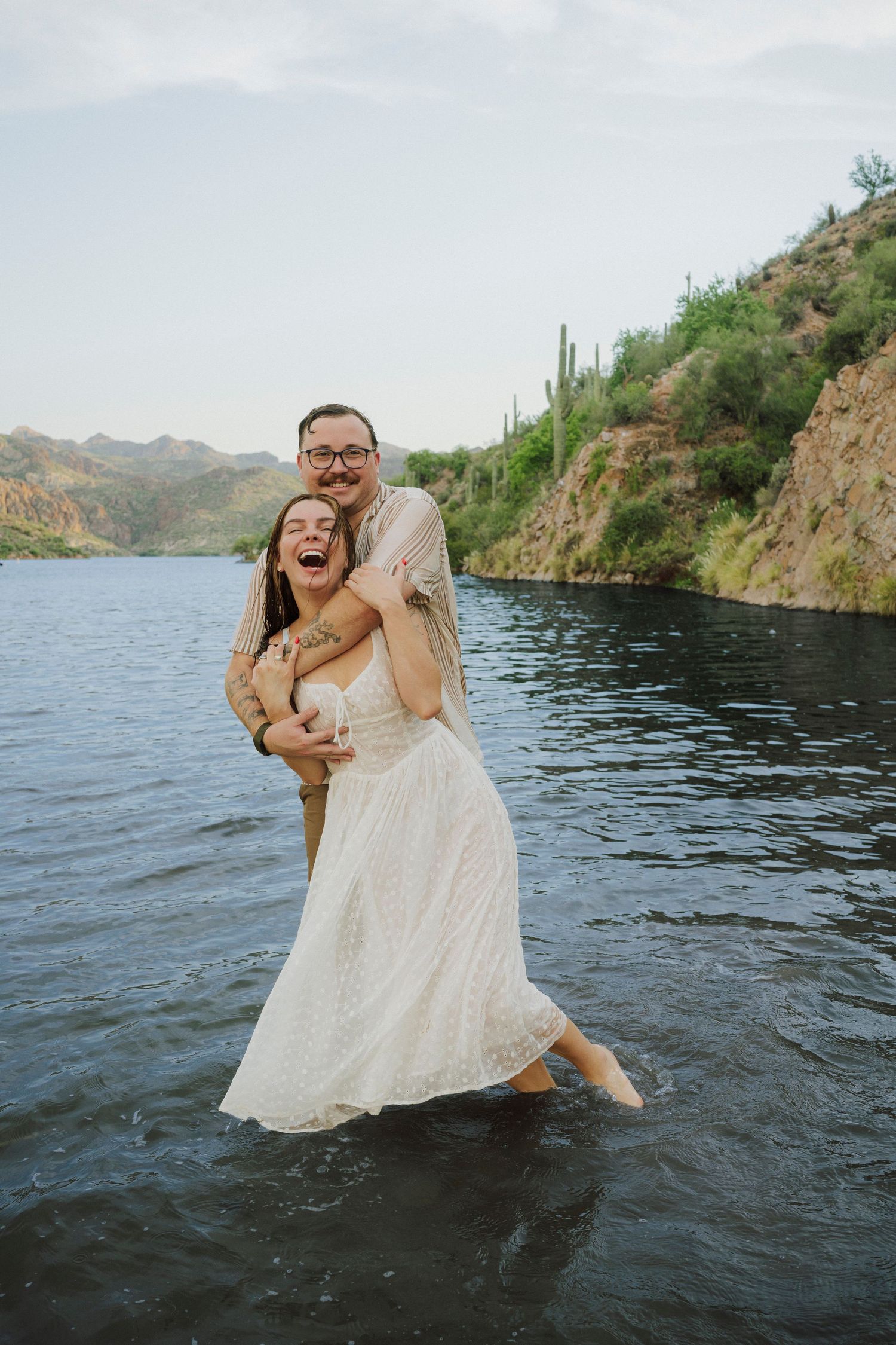 Joyful couple in wedding attire wading in a lake surrounded by desert mountains and saguaro cacti.