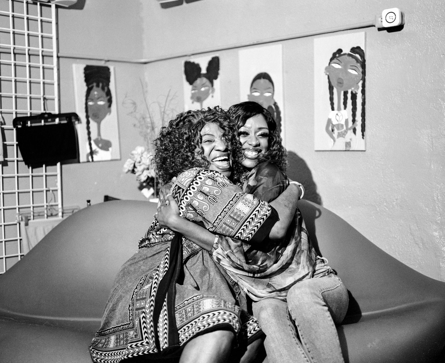 Black and white photo of two women hugging on couch with artwork on wall behind them.
