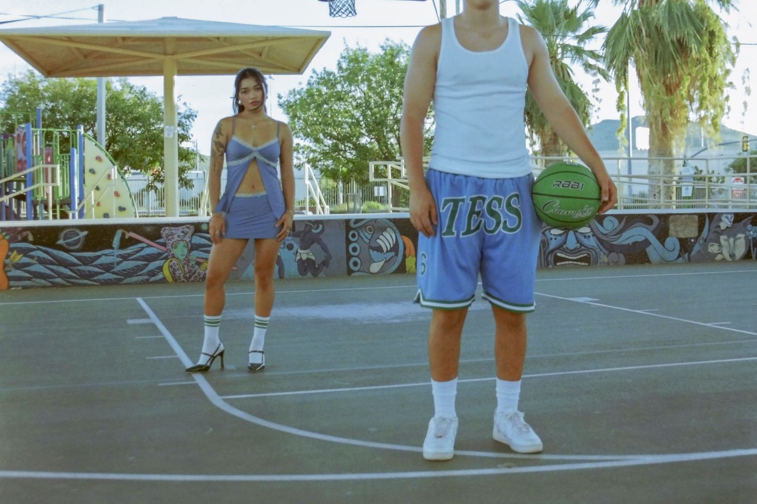 Young couple on outdoor basketball court, woman in blue crop set, man holding green basketball.