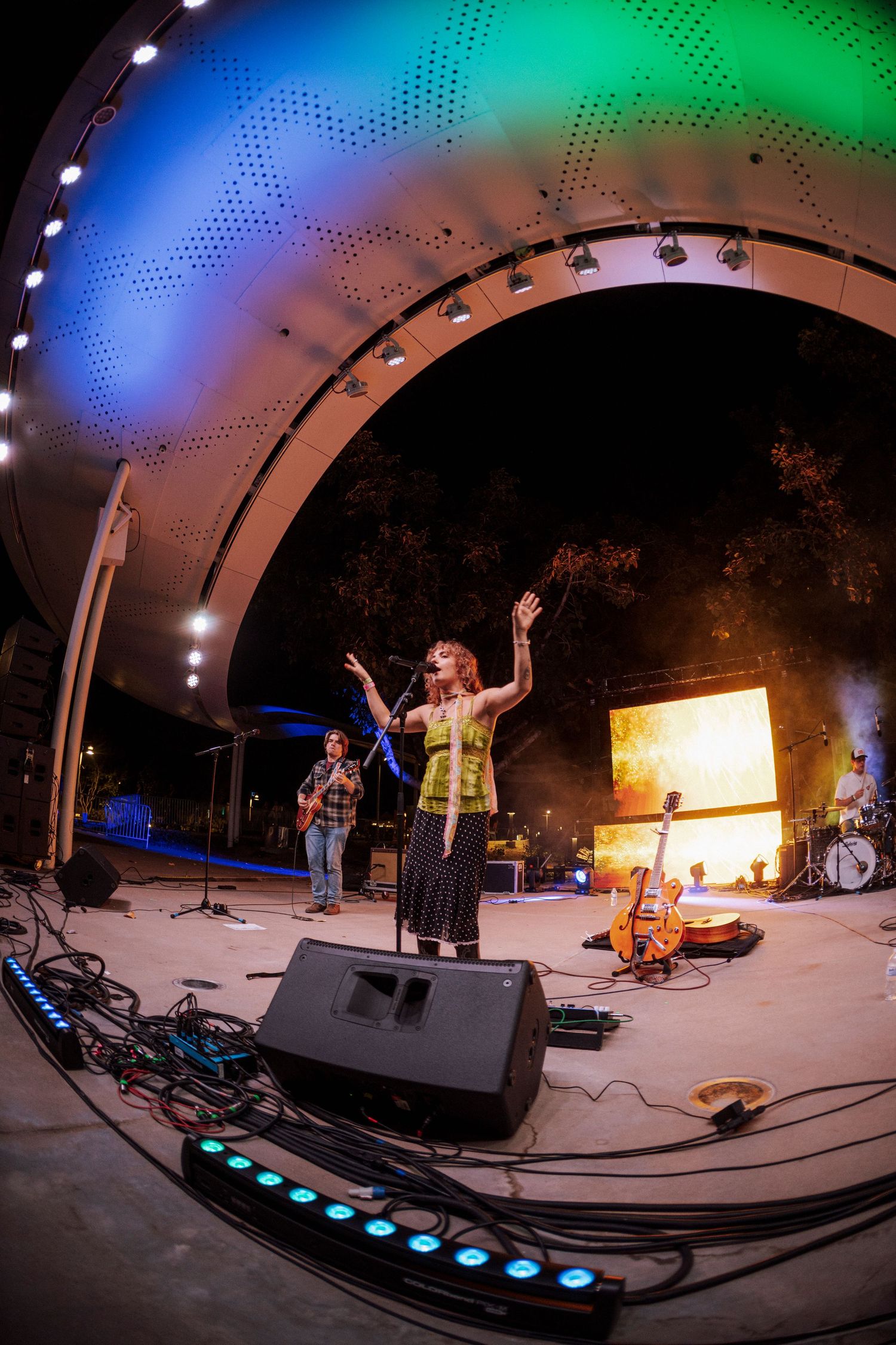 Female vocalist with arms raised performing on outdoor amphitheater stage with colorful arc lighting.