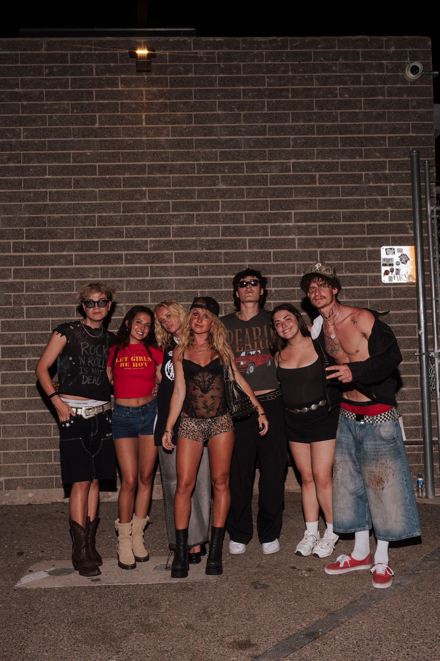 Group of young adults in edgy rock-inspired outfits posing together at night against a brick wall.