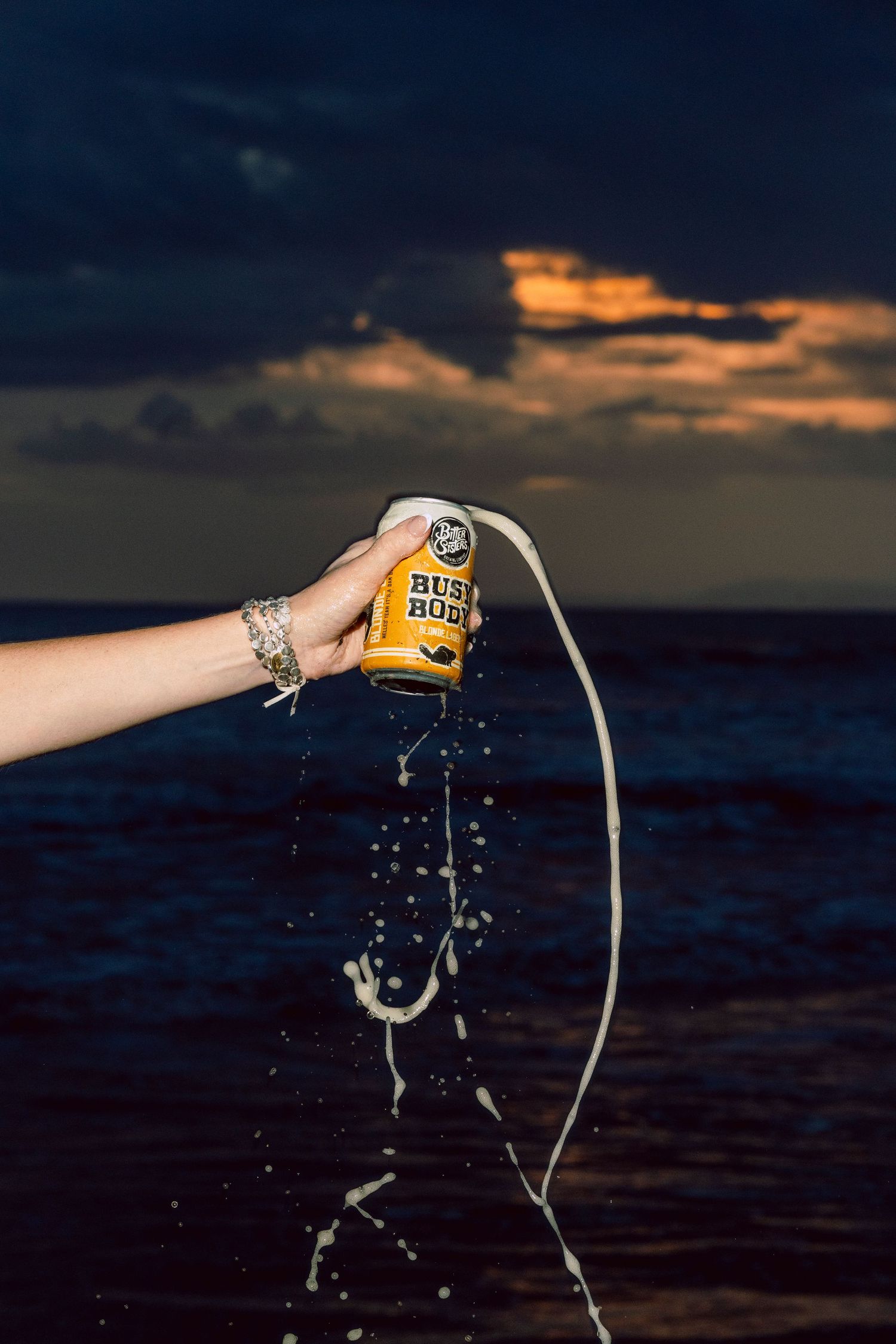 Hand holding a Rusty Bucket beer can spilling liquid at sunset over the ocean with dramatic cloudy sky.