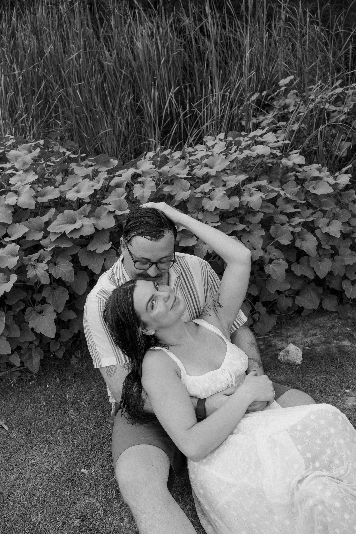 Black and white photo of a couple sharing a tender moment, sitting together in front of lush garden foliage.