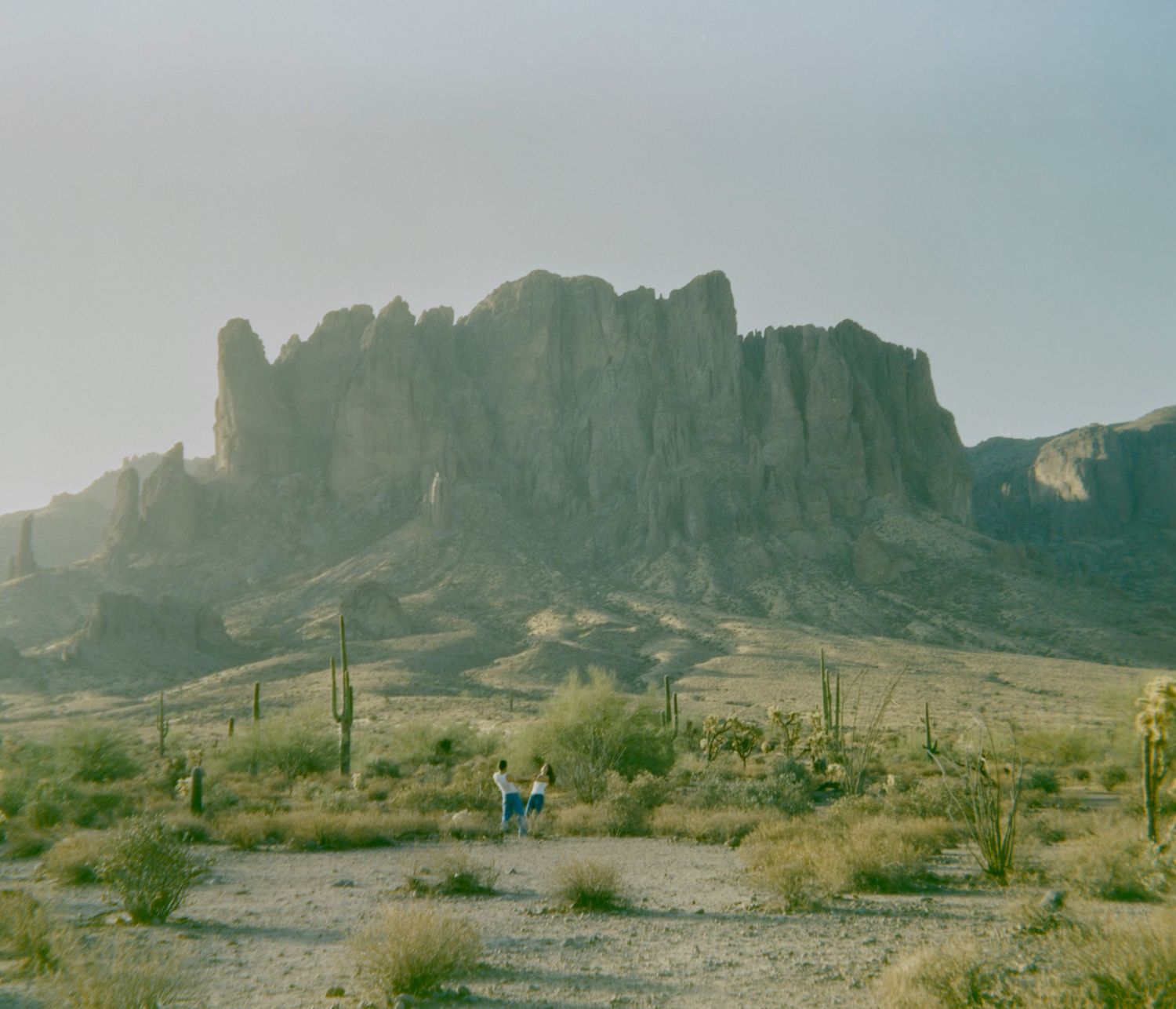 Dramatic desert landscape with towering rocky mountain, saguaro cacti, and two figures standing in the arid terrain below.