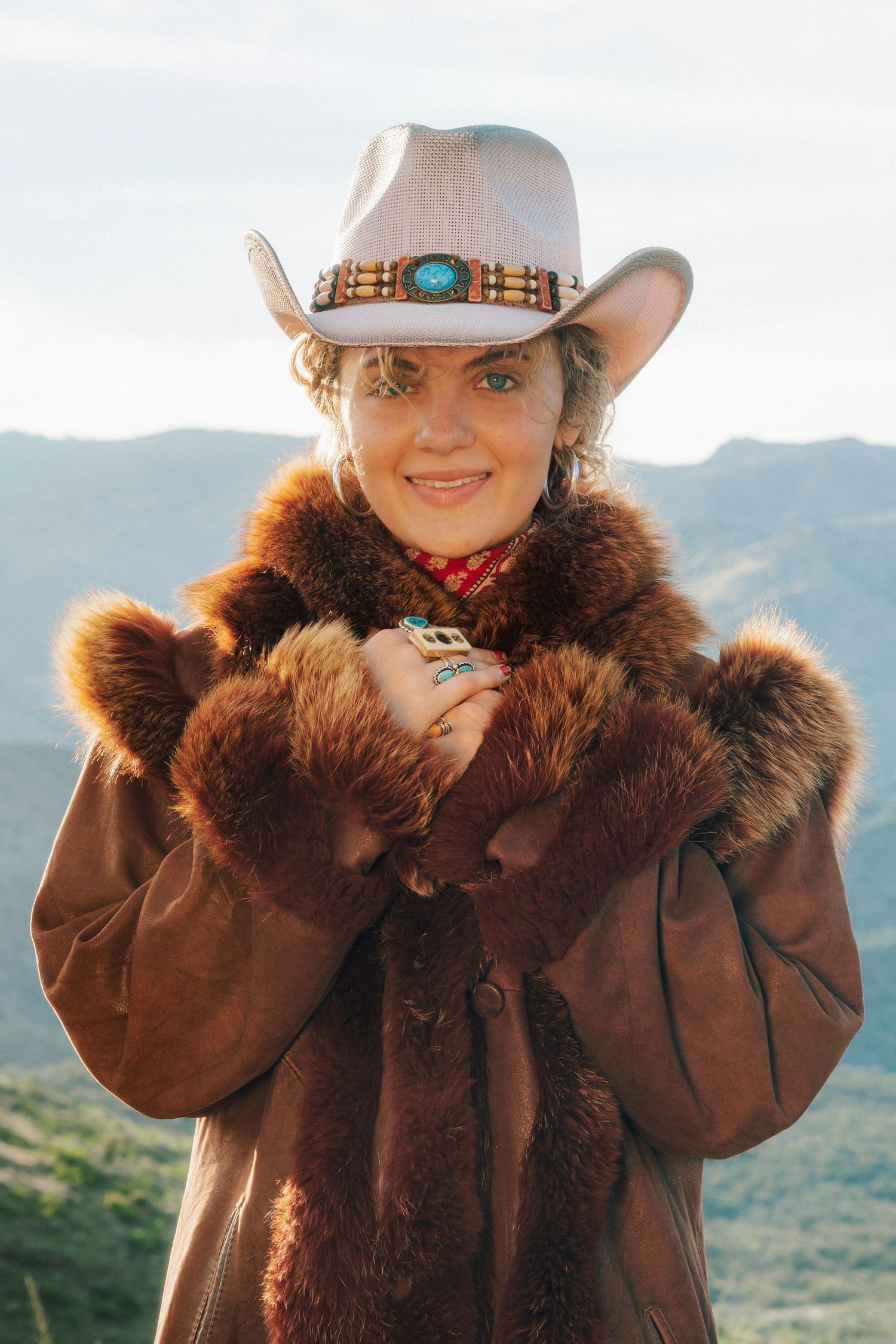 Smiling woman in turquoise-banded cowboy hat and brown fur coat poses with mountain backdrop.