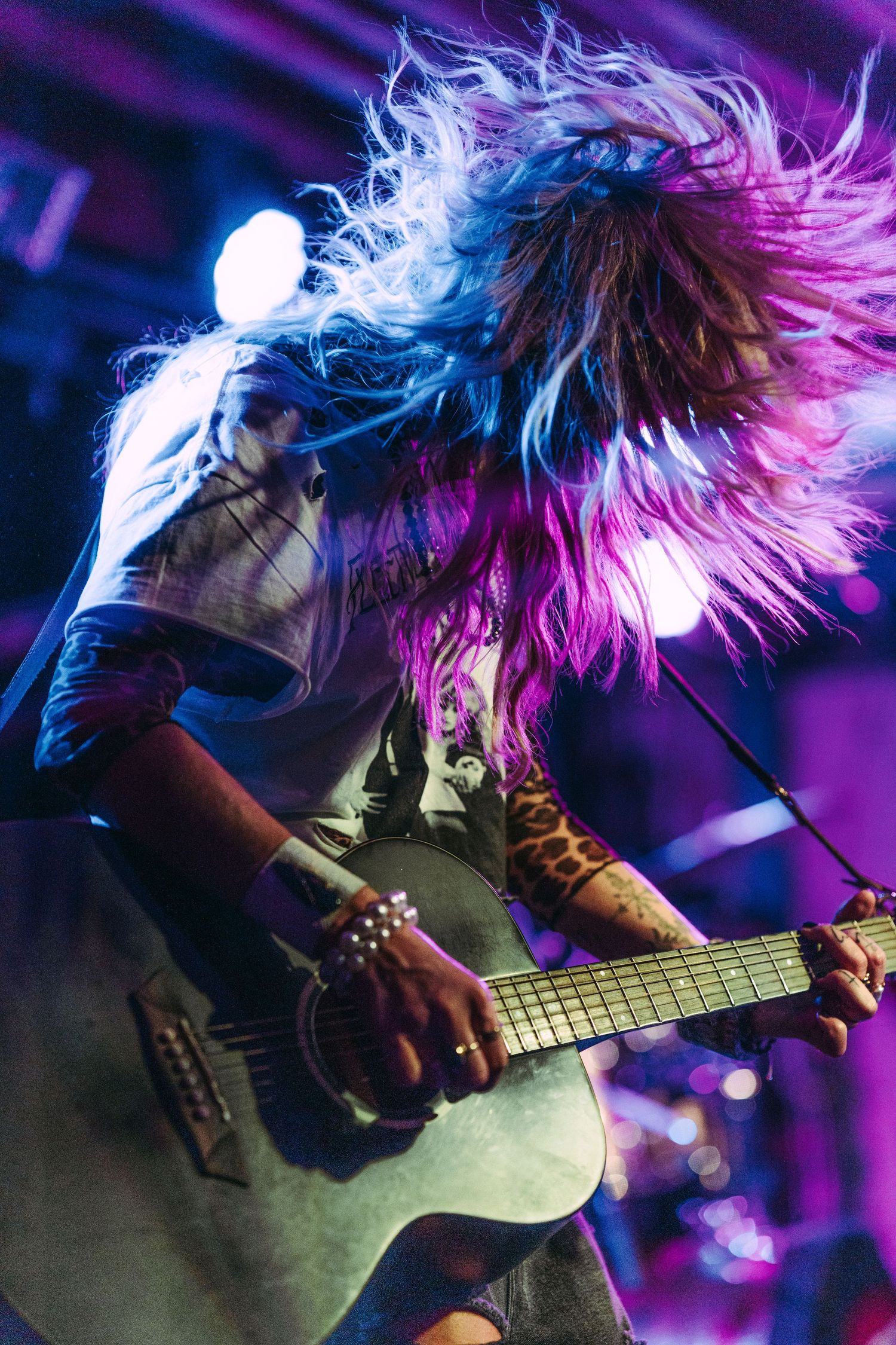 Guitarist headbanging with wild colorful hair while playing acoustic guitar under purple stage lights at concert.