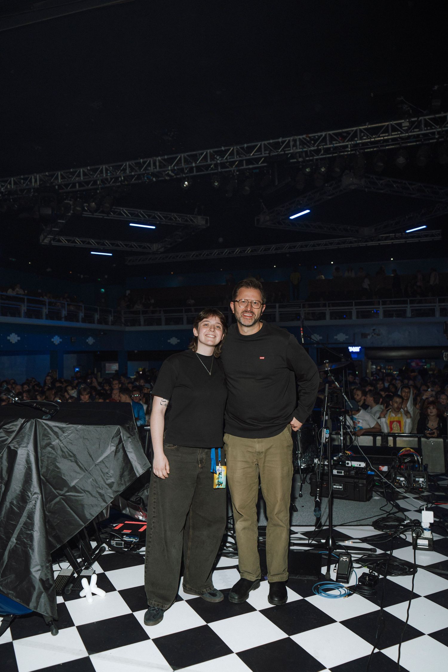 Two people posing together on a checkered floor stage at a large indoor concert venue with audience seating visible.