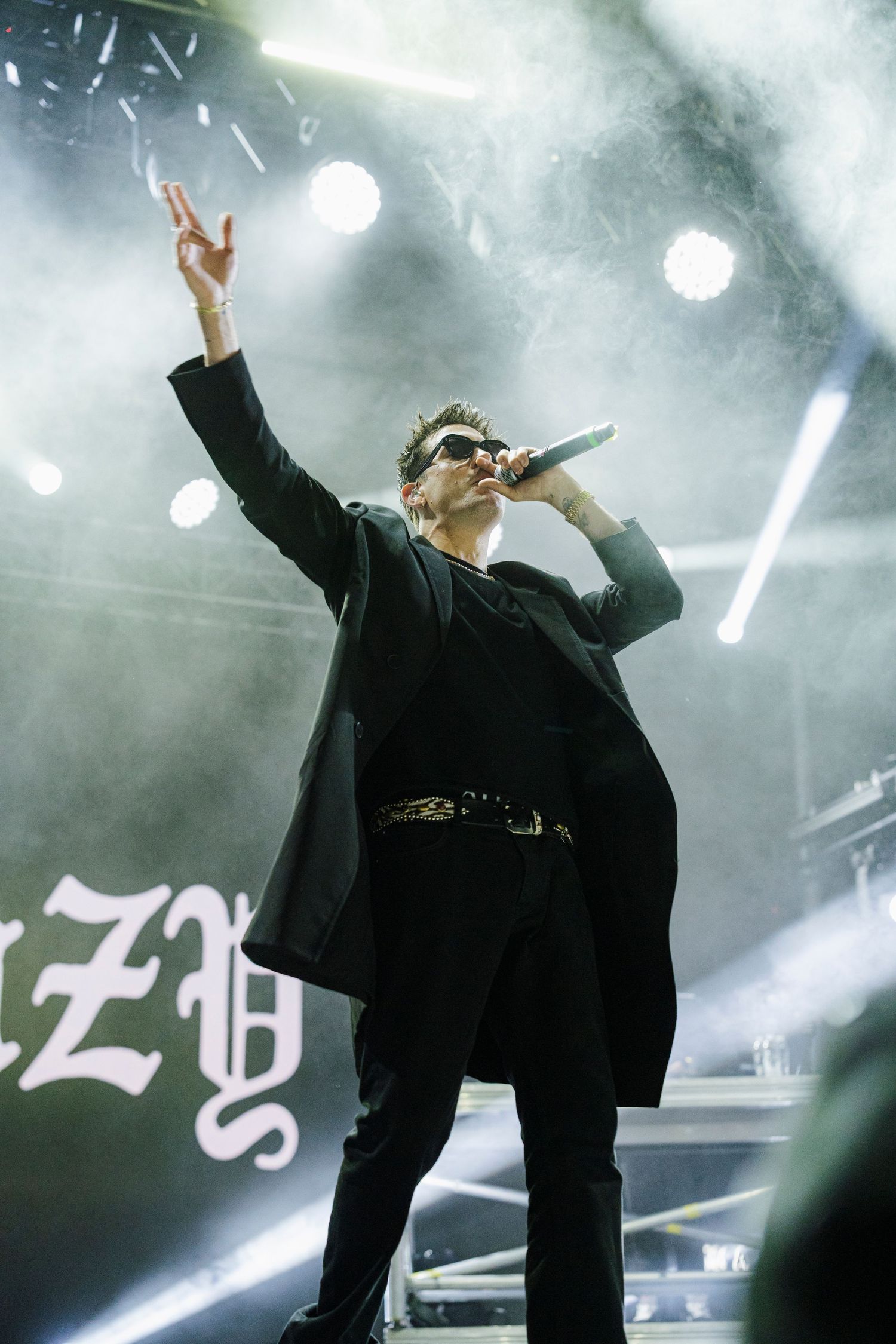 Male singer in black suit performing on stage with microphone raised hand and atmospheric fog lighting.