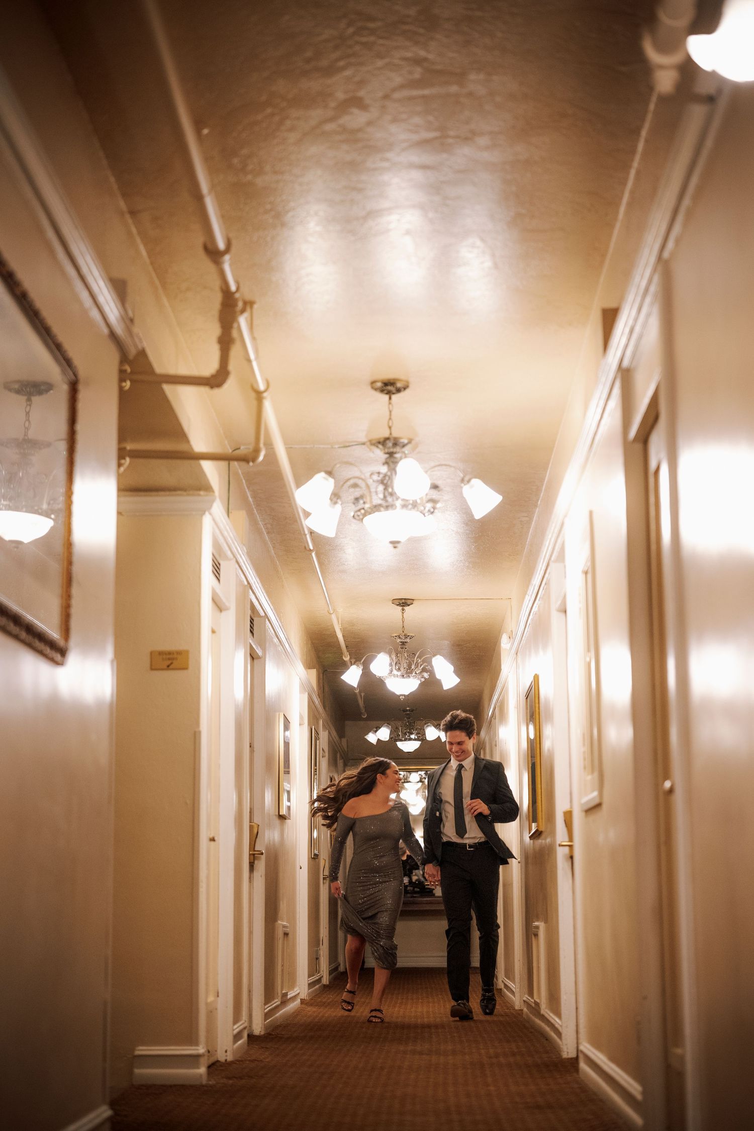 Couple running playfully down an ornate hotel corridor with glowing chandeliers.