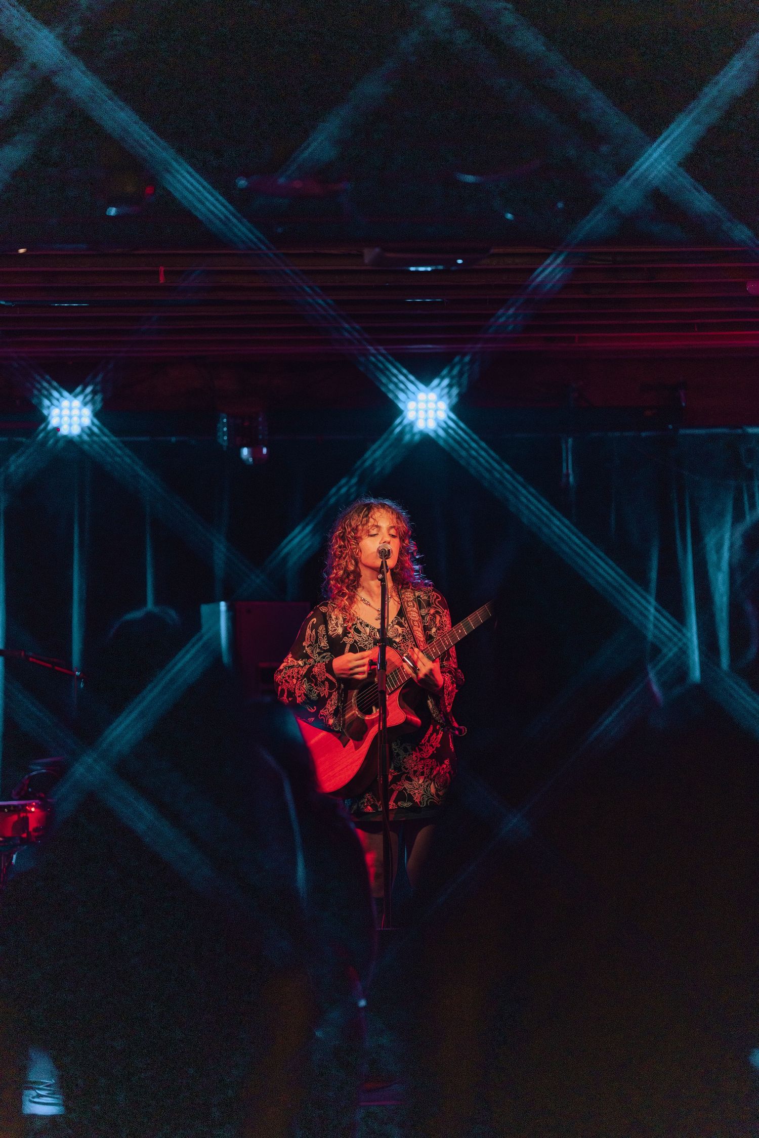 Curly-haired woman playing guitar on a dark stage framed by dramatic crossing beam lights.