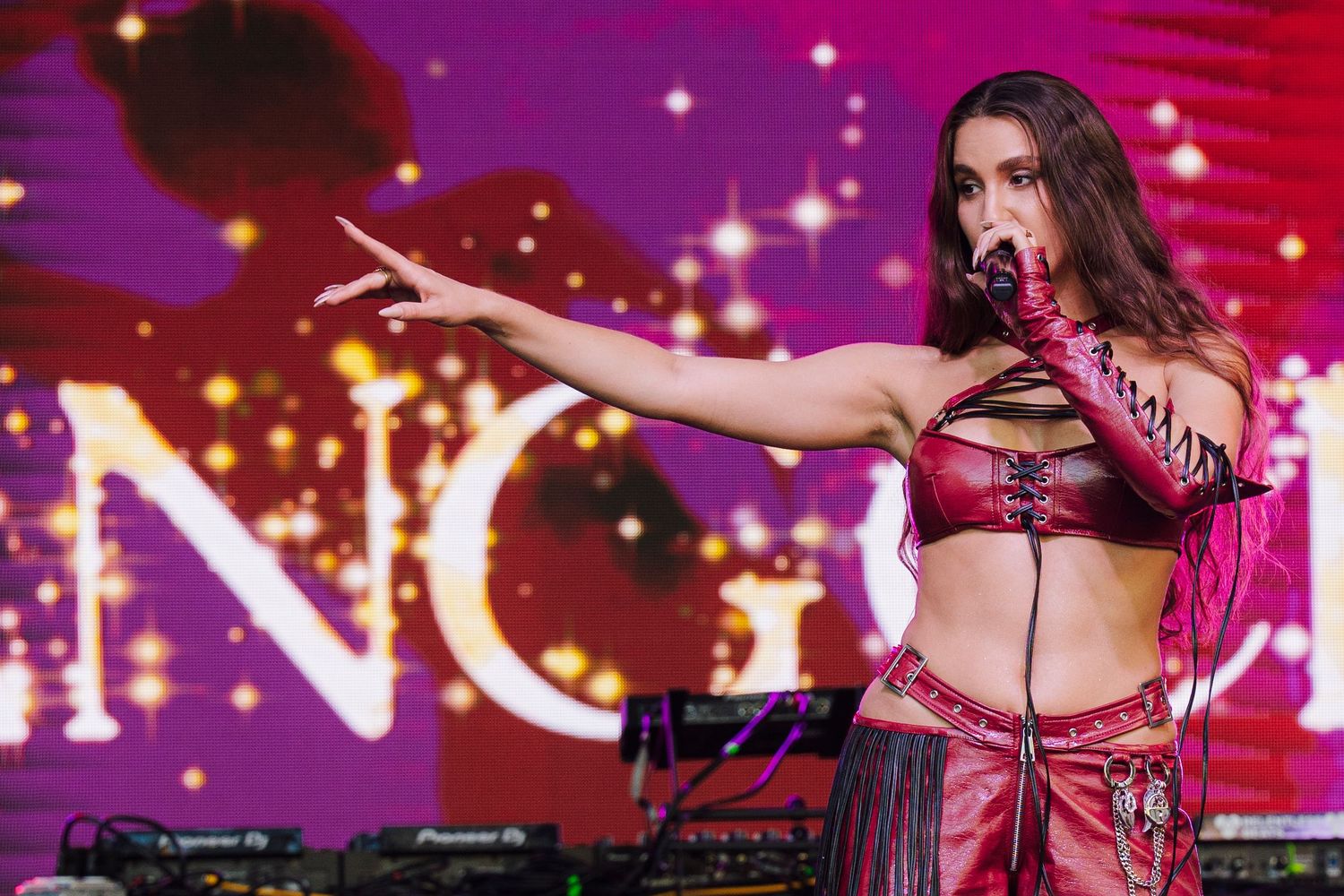Female performer in red leather outfit singing on stage with colorful purple backdrop at concert.