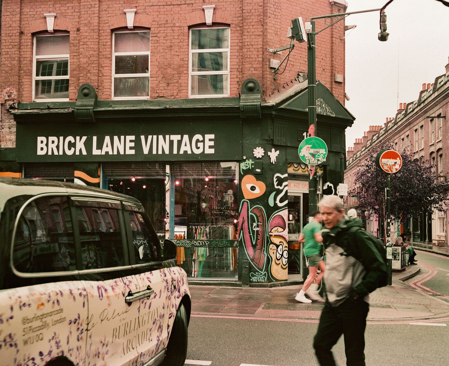 Brick Lane Vintage shop on a busy London street corner with a black cab and pedestrians passing by.