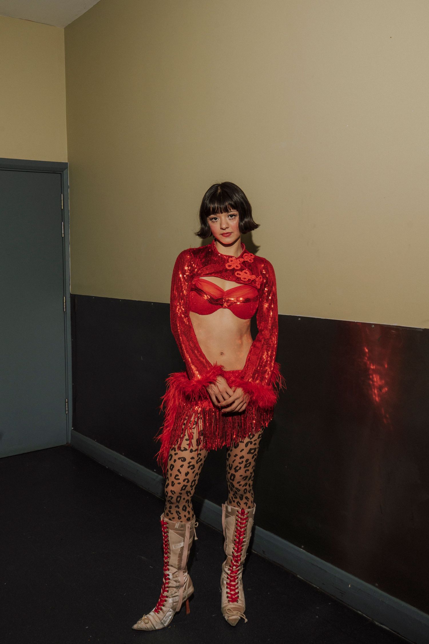 Woman in red sequin crop top, leopard print tights, and lace-up heels poses in a hallway.