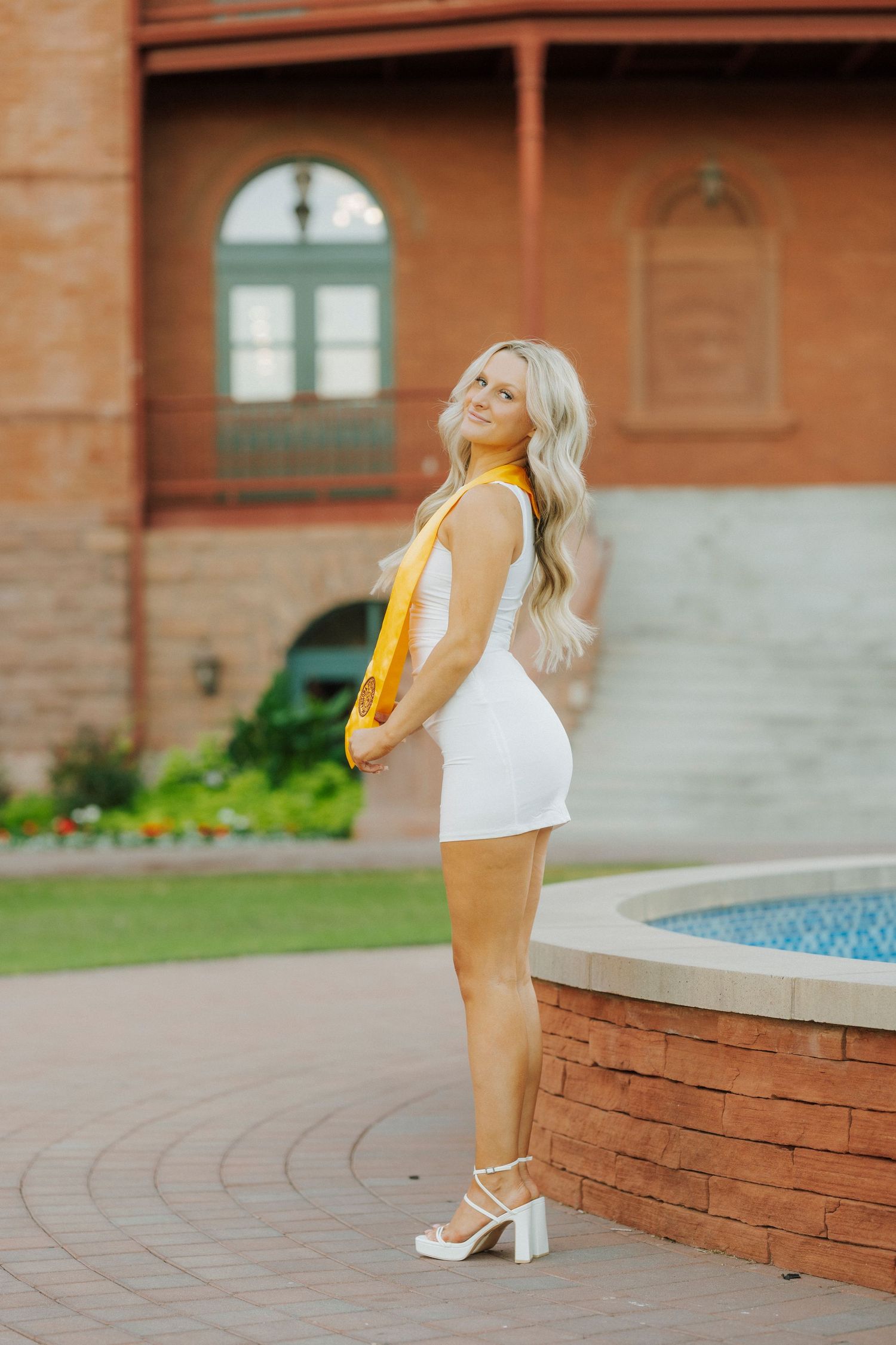 Blonde woman in white mini dress and yellow sash posing by fountain outside brick building.