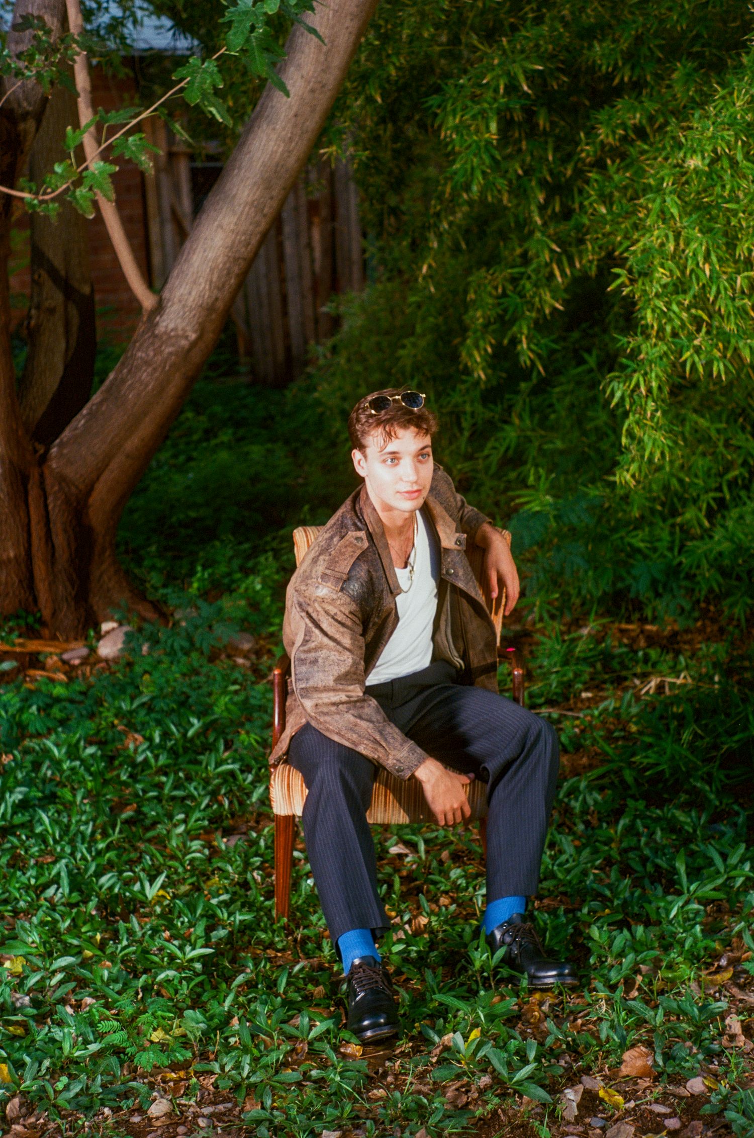 Young man in brown jacket and blue socks sits on chair surrounded by lush greenery.
