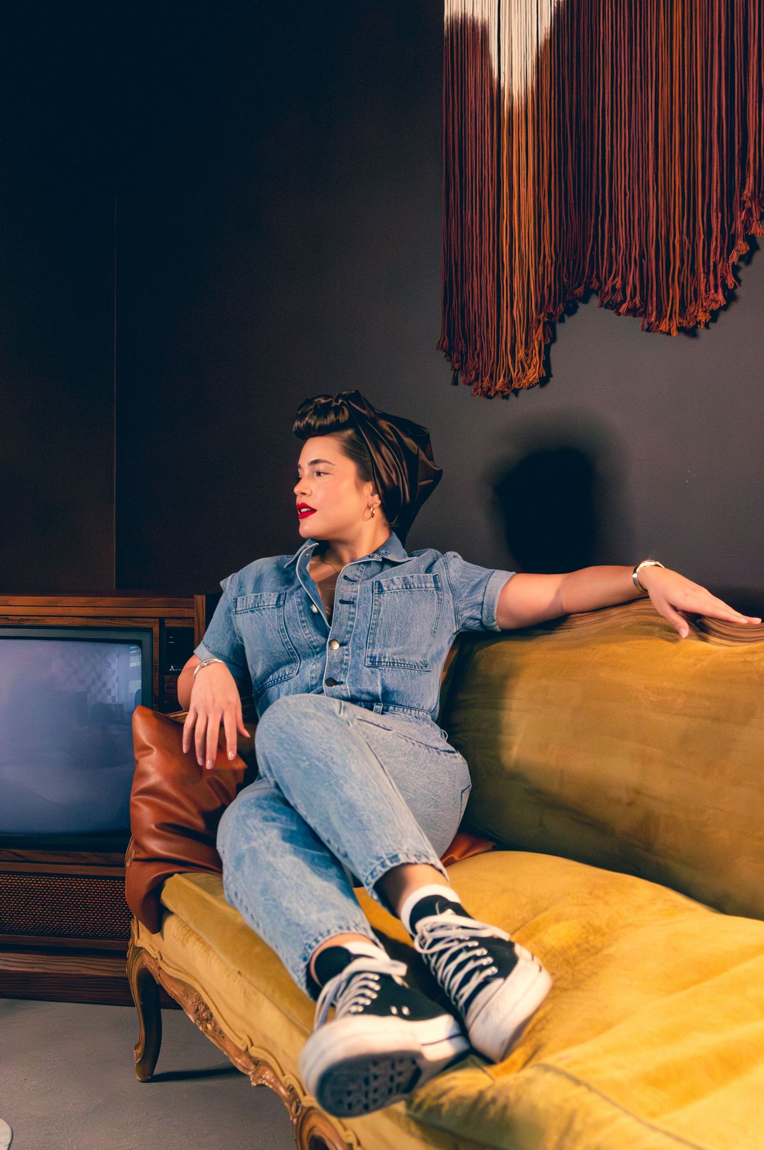 Woman in denim jumpsuit relaxing on vintage gold velvet sofa next to retro television set.