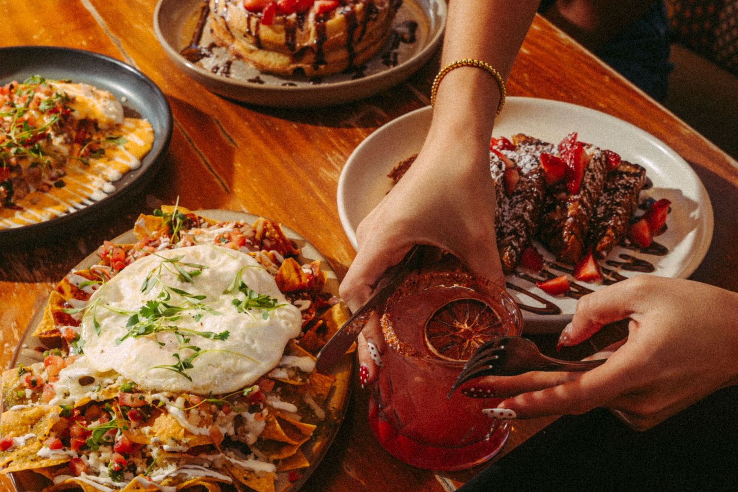 Hands reaching for a cocktail beside loaded nachos and chocolate cake on a wooden table.