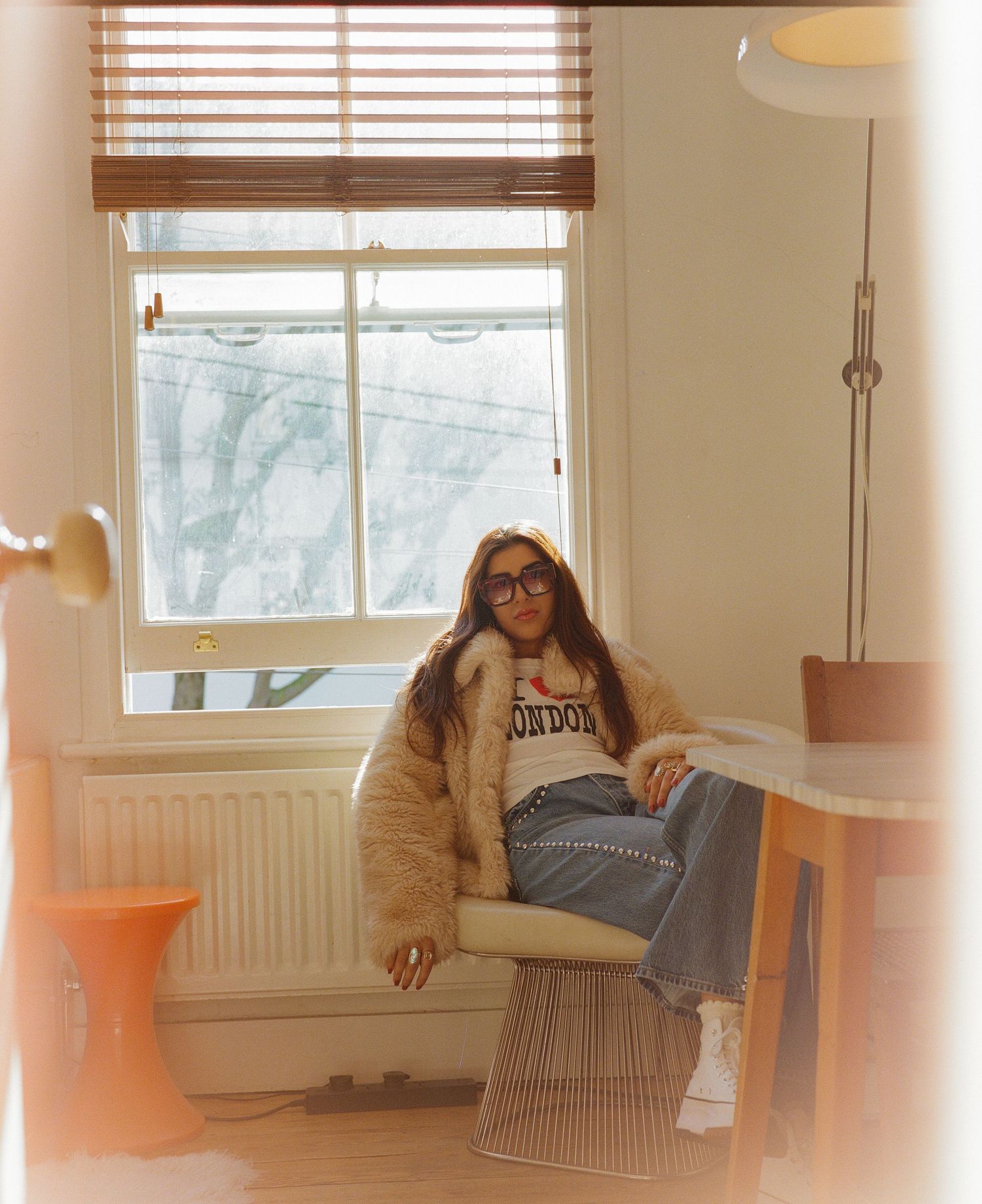 Woman in fuzzy coat and jeans sitting by window with London shirt in cozy interior.