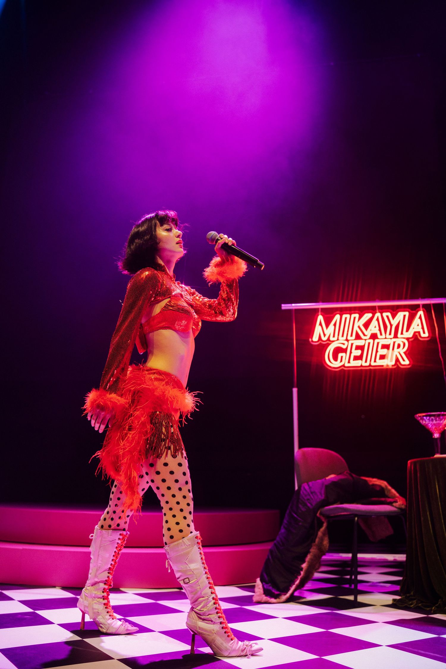 Mikayla Geier performs on stage in a red feathered costume with polka dot tights on a checkered floor under purple lights.