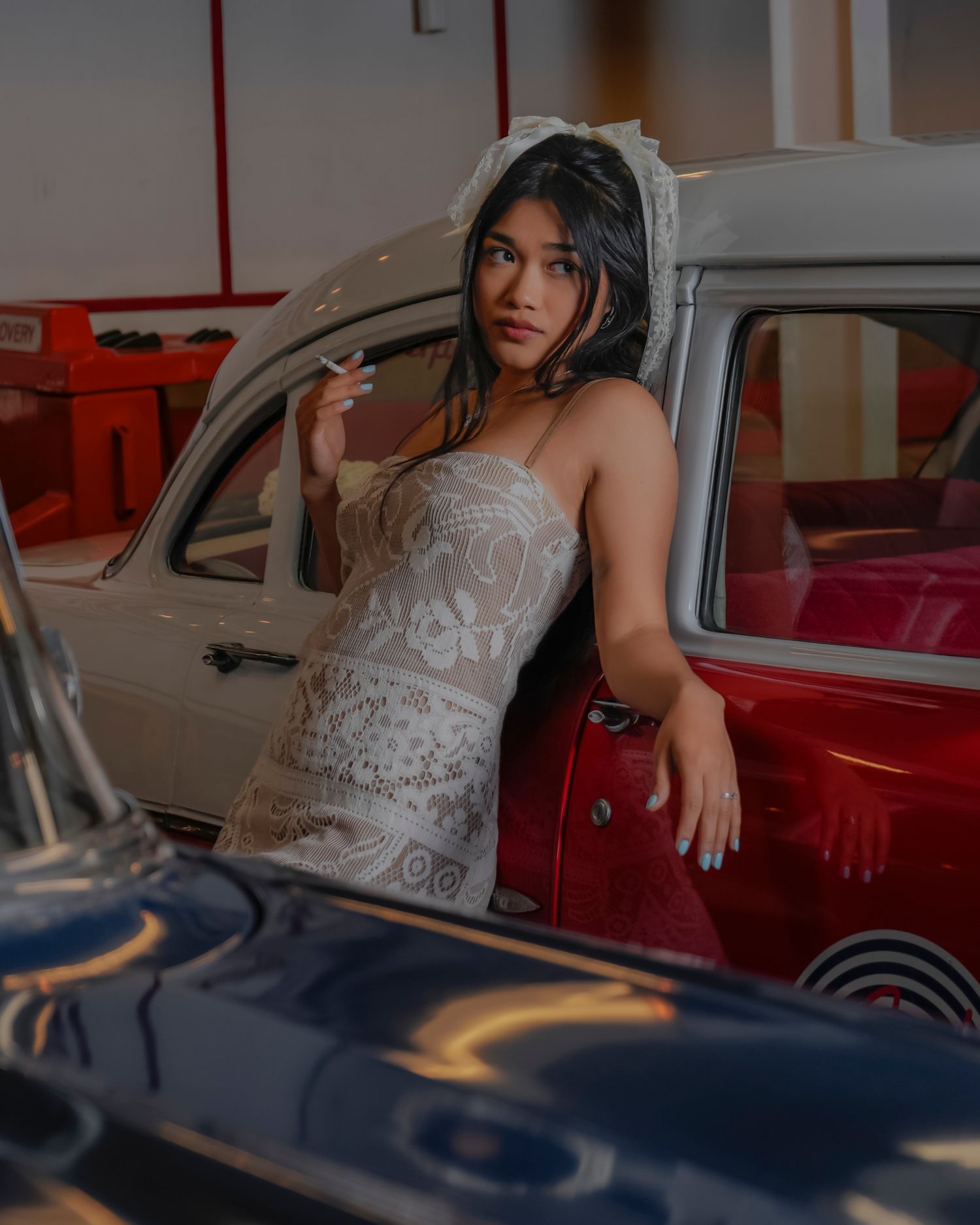 Young woman in lace mini dress and bridal veil poses against vintage red and white cars indoors.