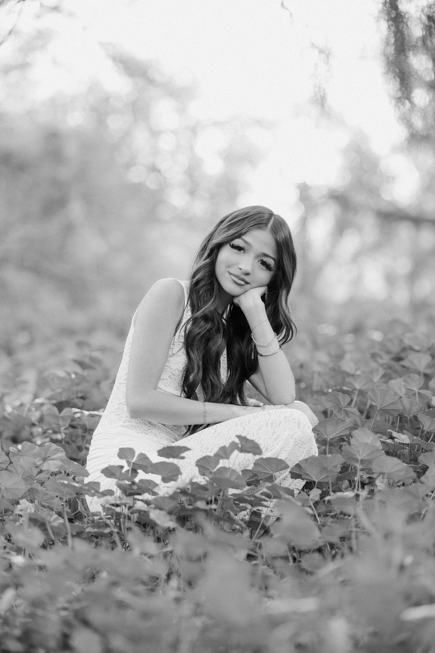 Young woman in white dress sits smiling among plants in black and white portrait.