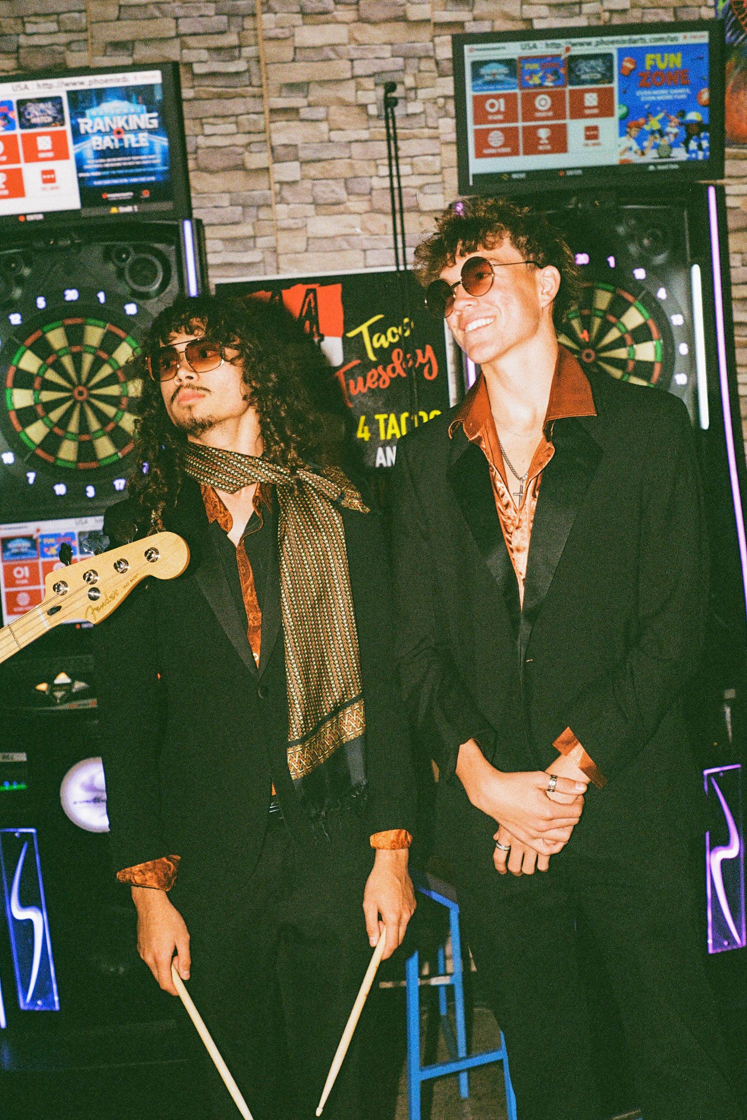 Two stylish young musicians in black suits with sunglasses posing in a bar near arcade games and dartboard.
