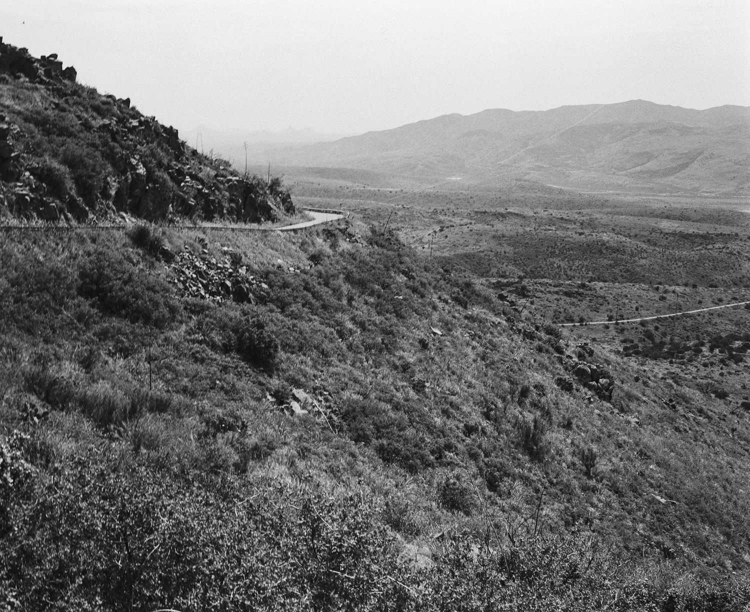 Black and white aerial view of a winding mountain road through a vast valley landscape.