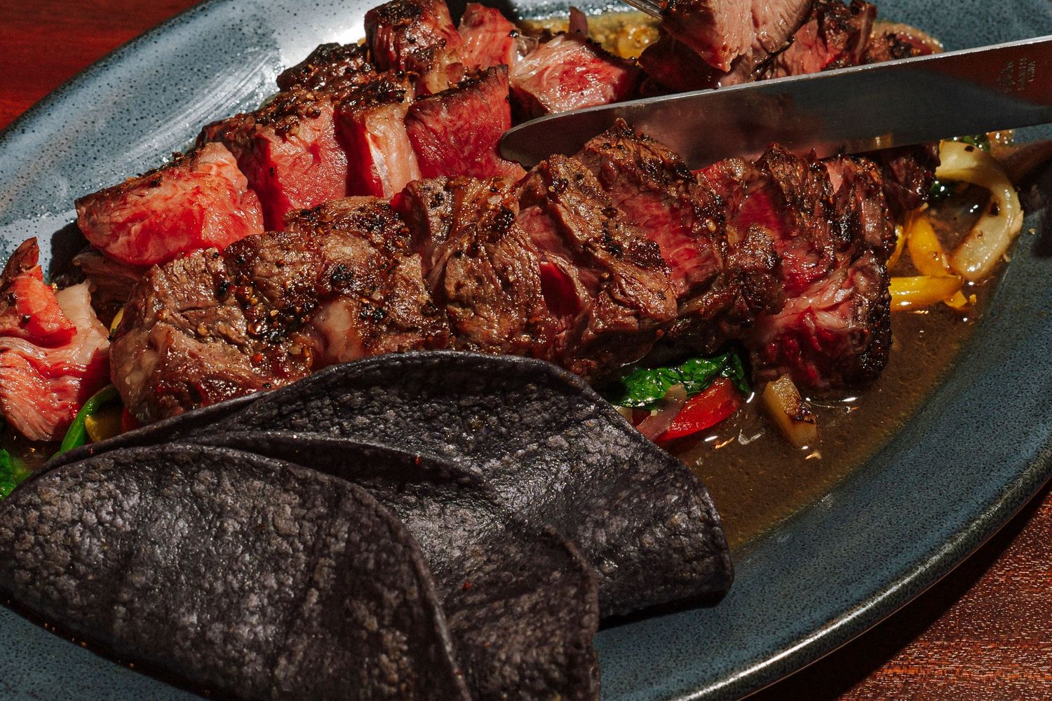 Grilled skewered meat served on a blue plate with colorful vegetables and black tortillas.