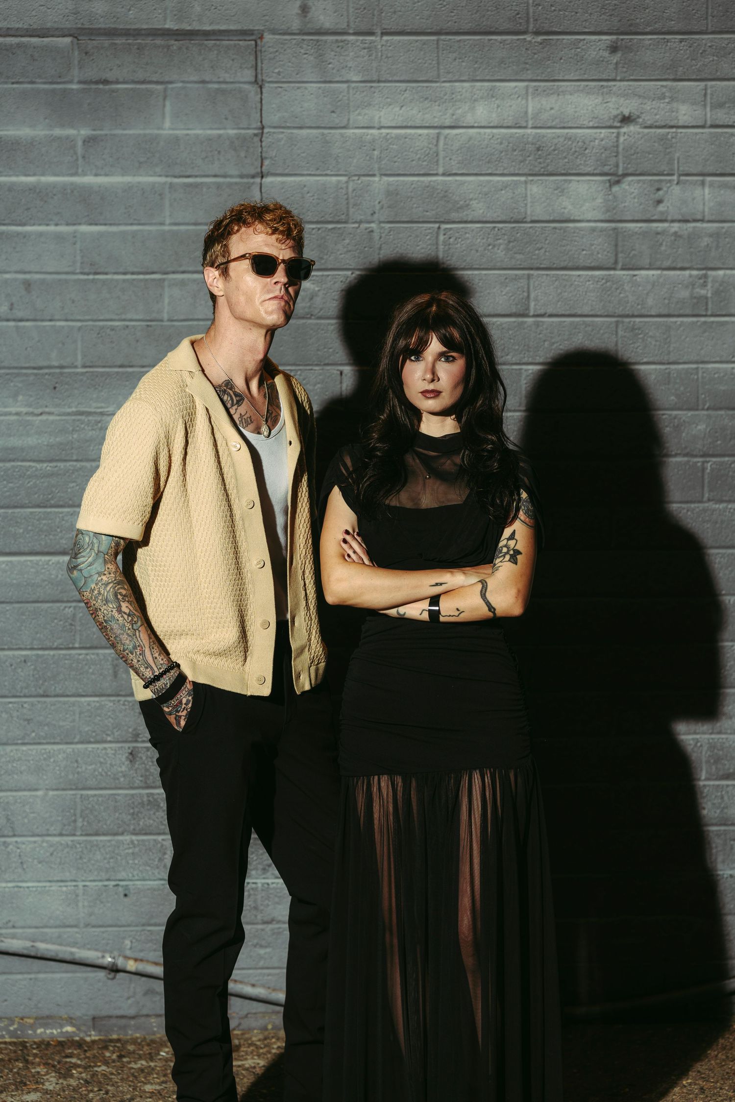 Tattooed couple posing against a gray wall, man in cardigan, woman in black dress.