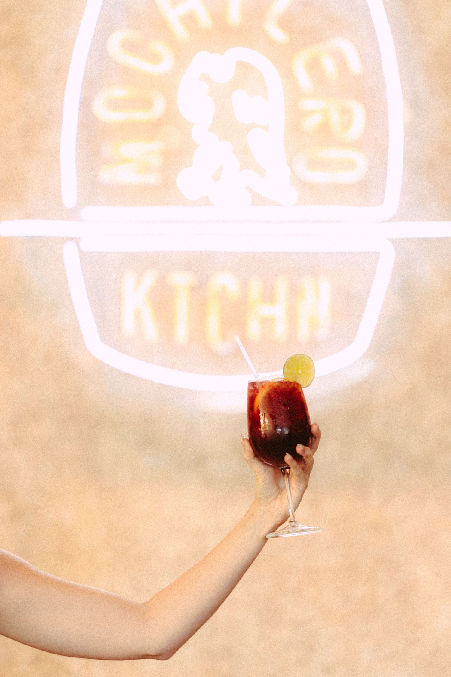 Hand holding a red cocktail with lime garnish in front of Mochilero KTCHN neon sign.