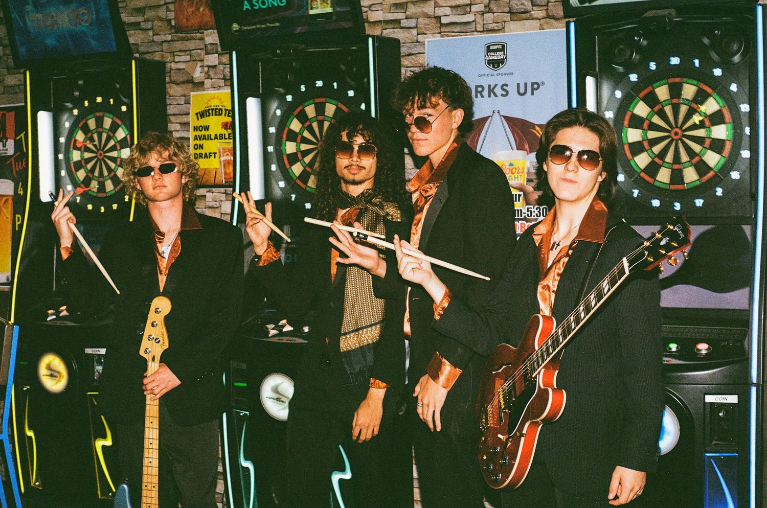Five musicians in black outfits pose with instruments near a dartboard in a bar setting.