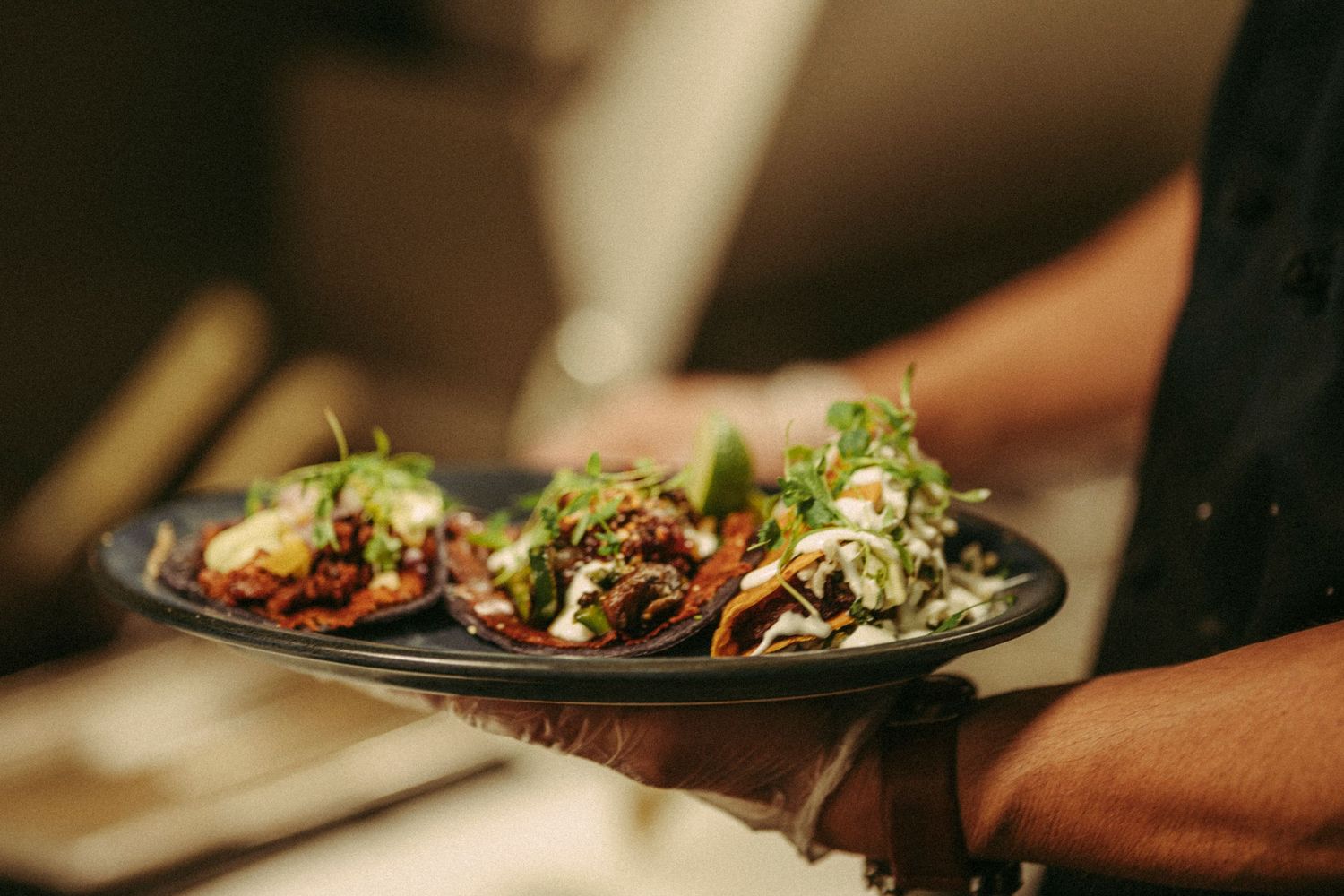 Server carrying a black plate of assorted gourmet tacos with fresh toppings.
