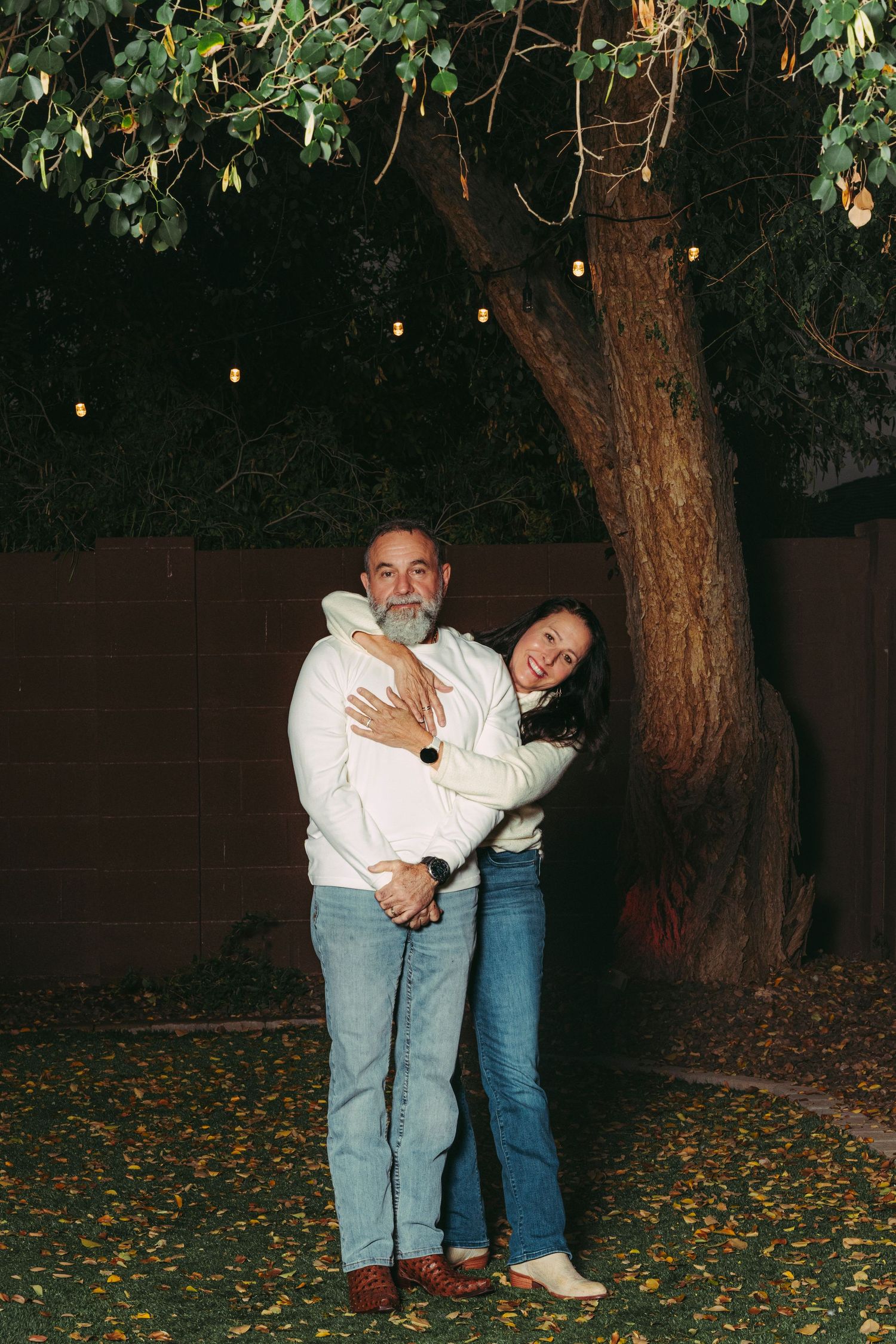 Couple embracing under a large tree with string lights at night, wearing casual jeans and white tops.