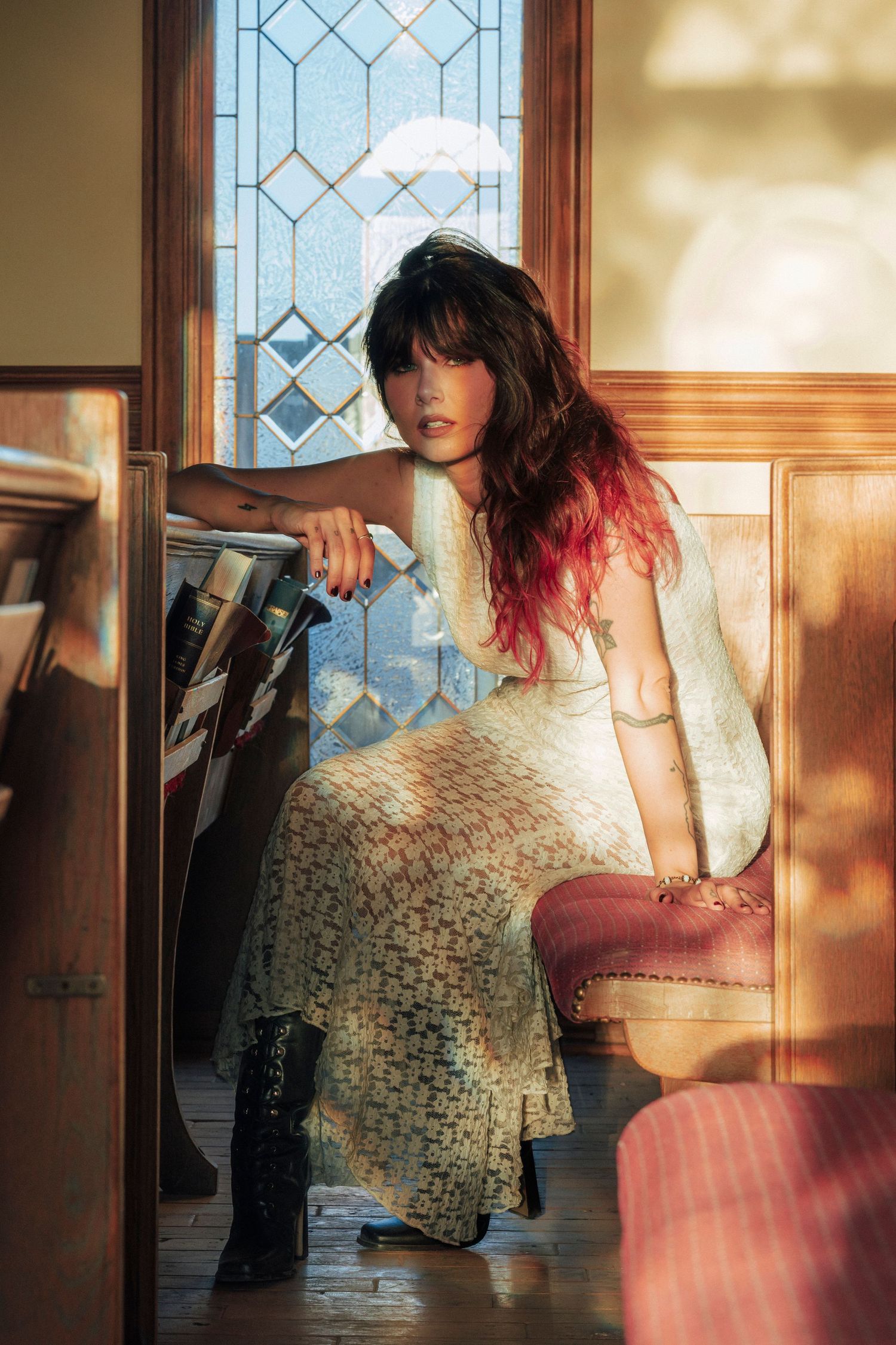 Woman in lace dress and black boots sits by stained glass window in warm sunlit vintage interior.