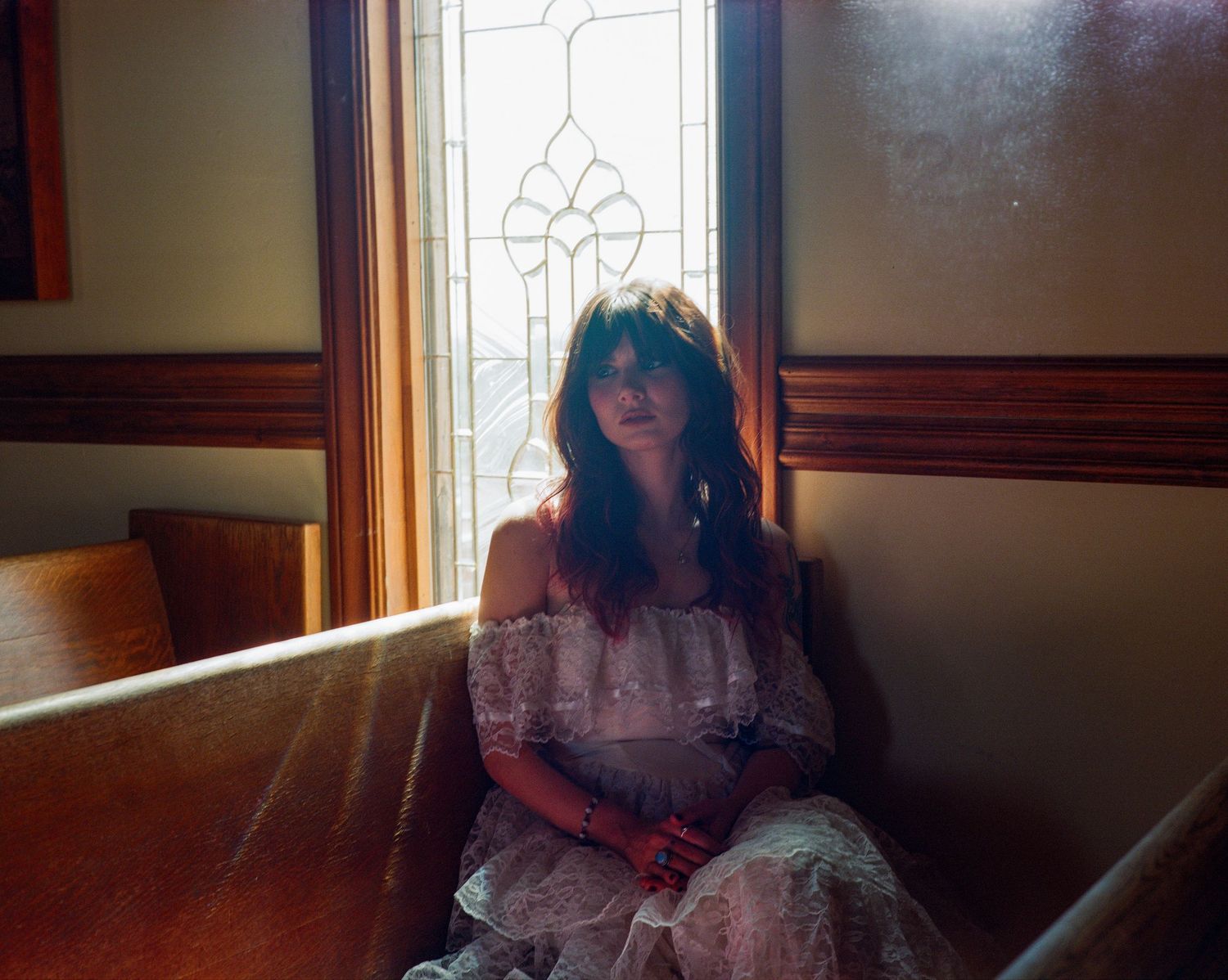 Young woman in white lace off-shoulder dress sitting in a church pew near a stained glass window. From 'Dead Ends' music video