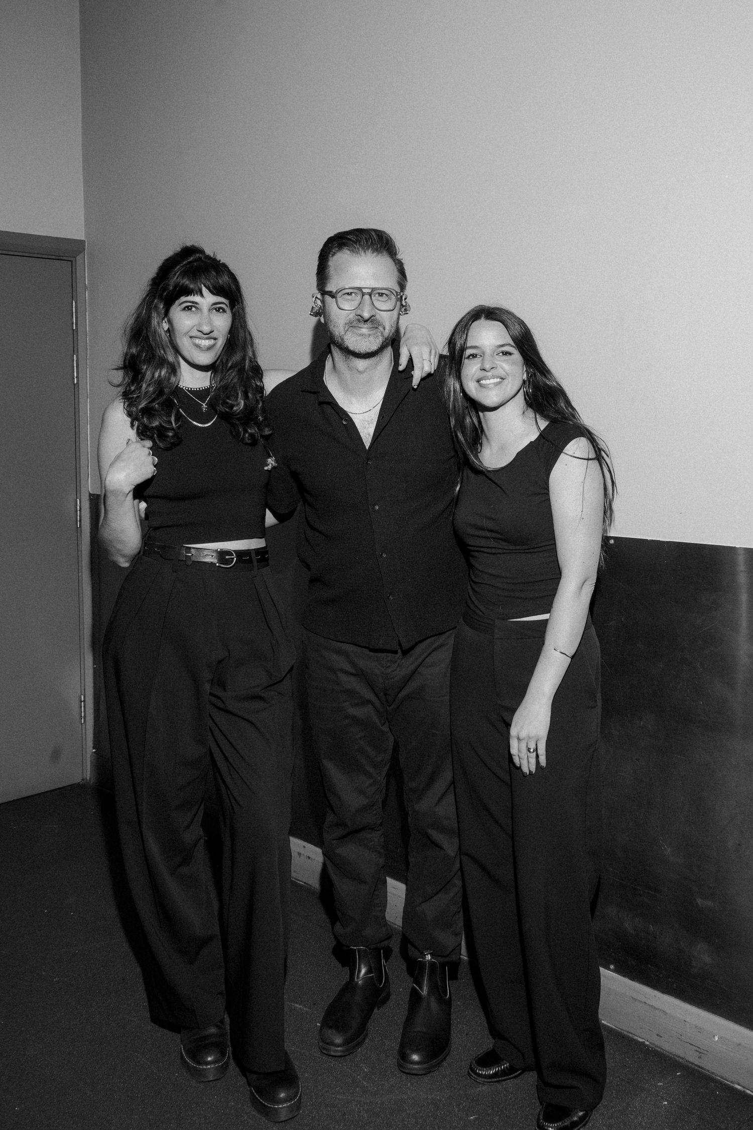 Three people dressed in black posing together for a black and white photo indoors.