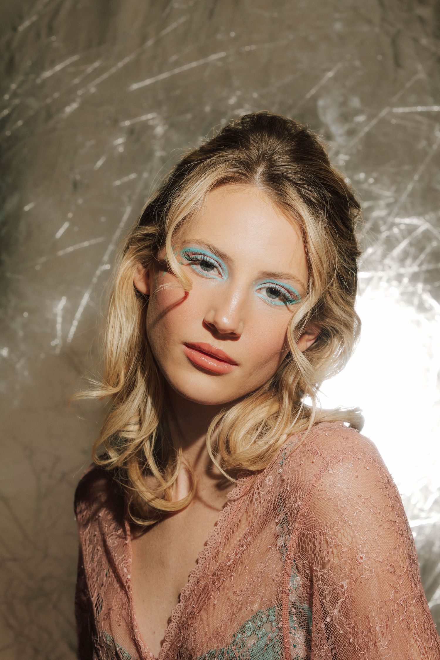 Blonde woman with blue glitter eye makeup in pink lace top poses against metallic background.