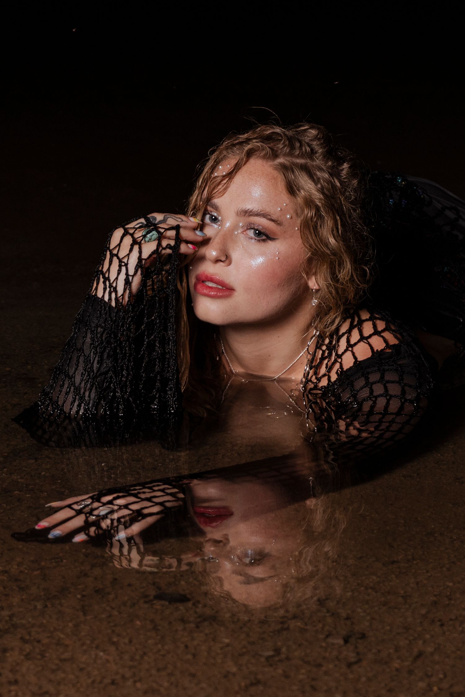 Young woman in black fishnet sleeves lying in shallow water at night with reflection visible.