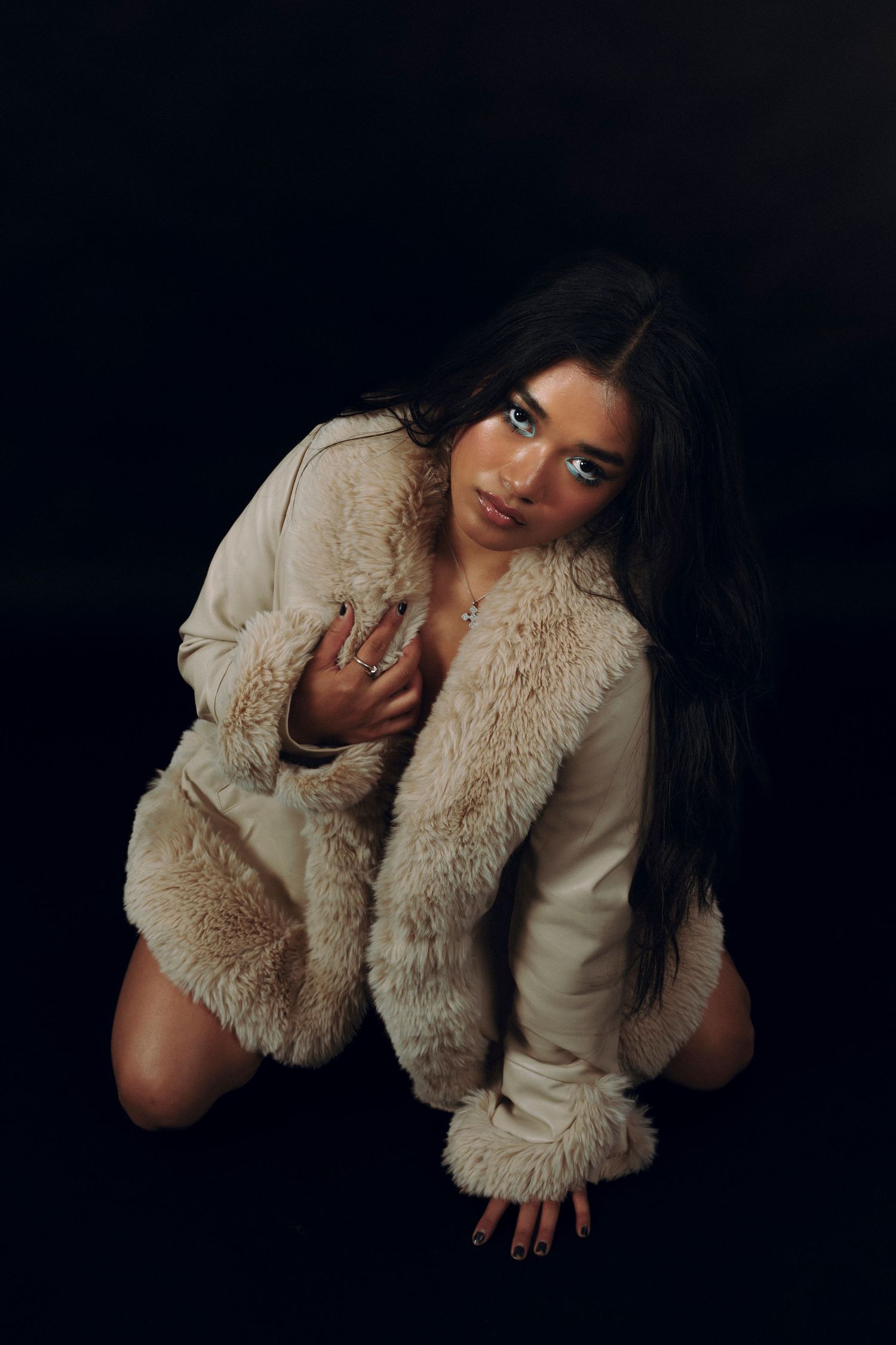 A young woman in a cream faux fur coat crouches on a dark studio floor, gazing at the camera with long dark hair.