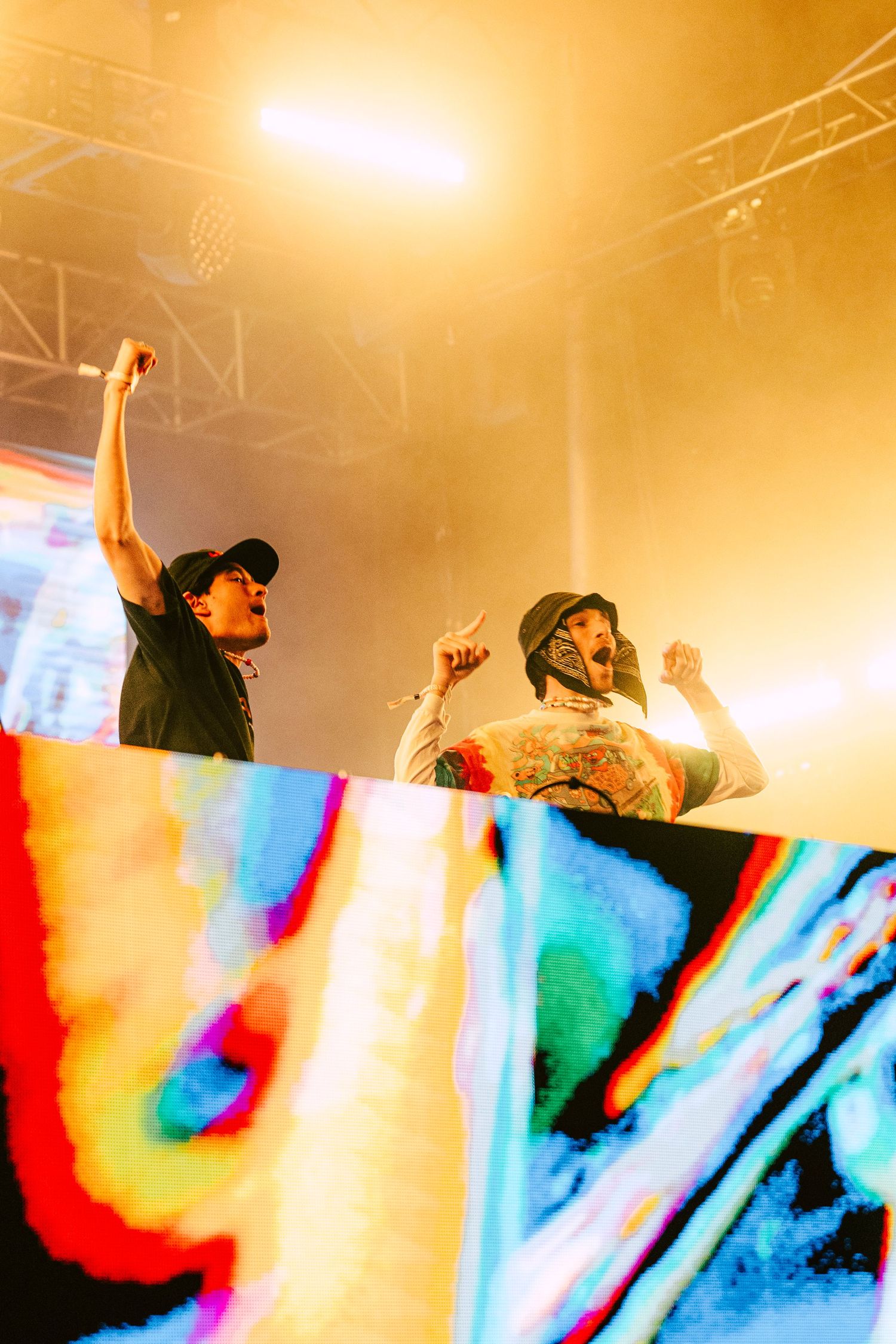 Two DJs performing on a colorful illuminated stage, raising their fists in the air under golden festival lights. Levity at Dusk 2025.