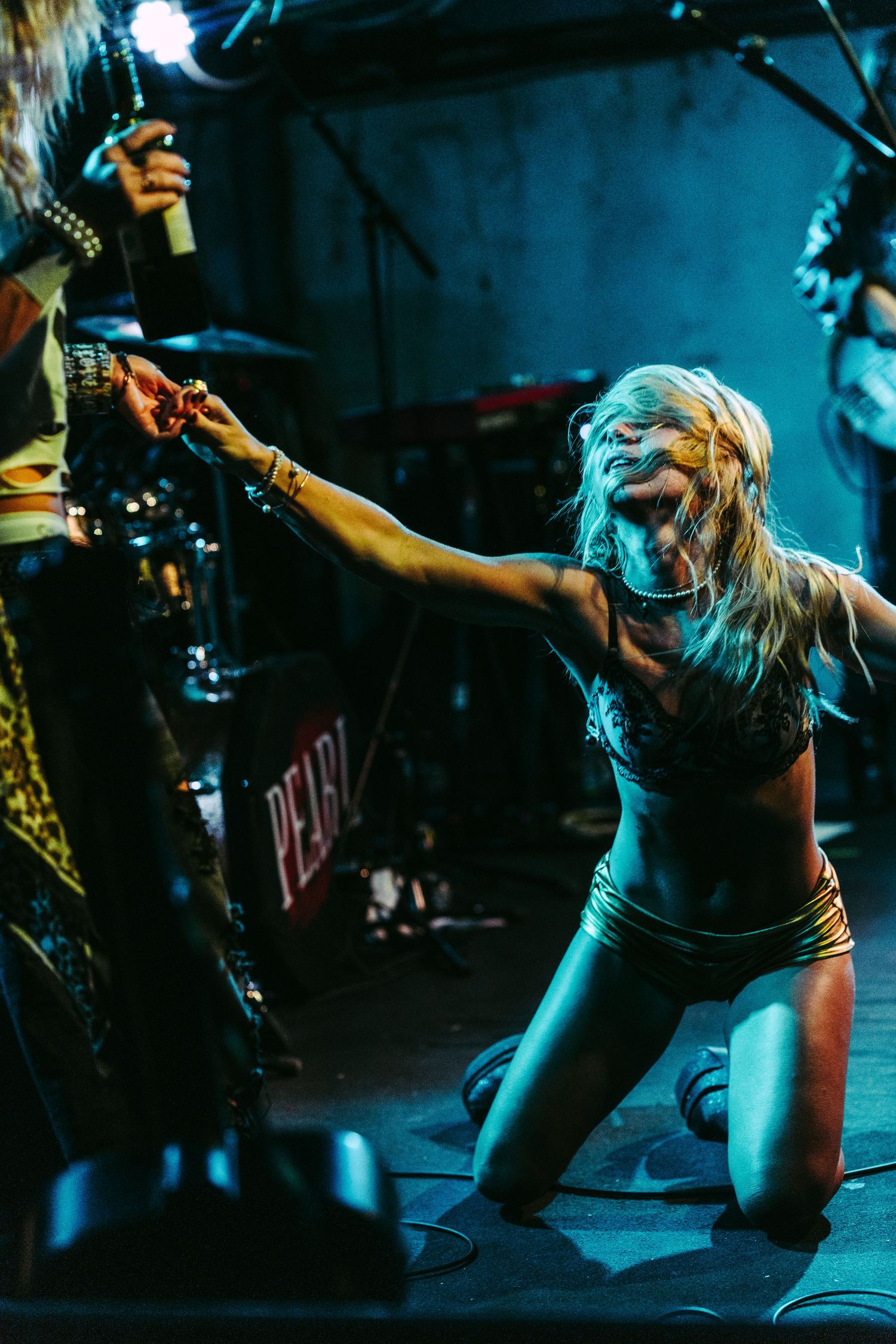 Blonde female rock performer kneeling on stage reaching toward another musician during an energetic live concert.