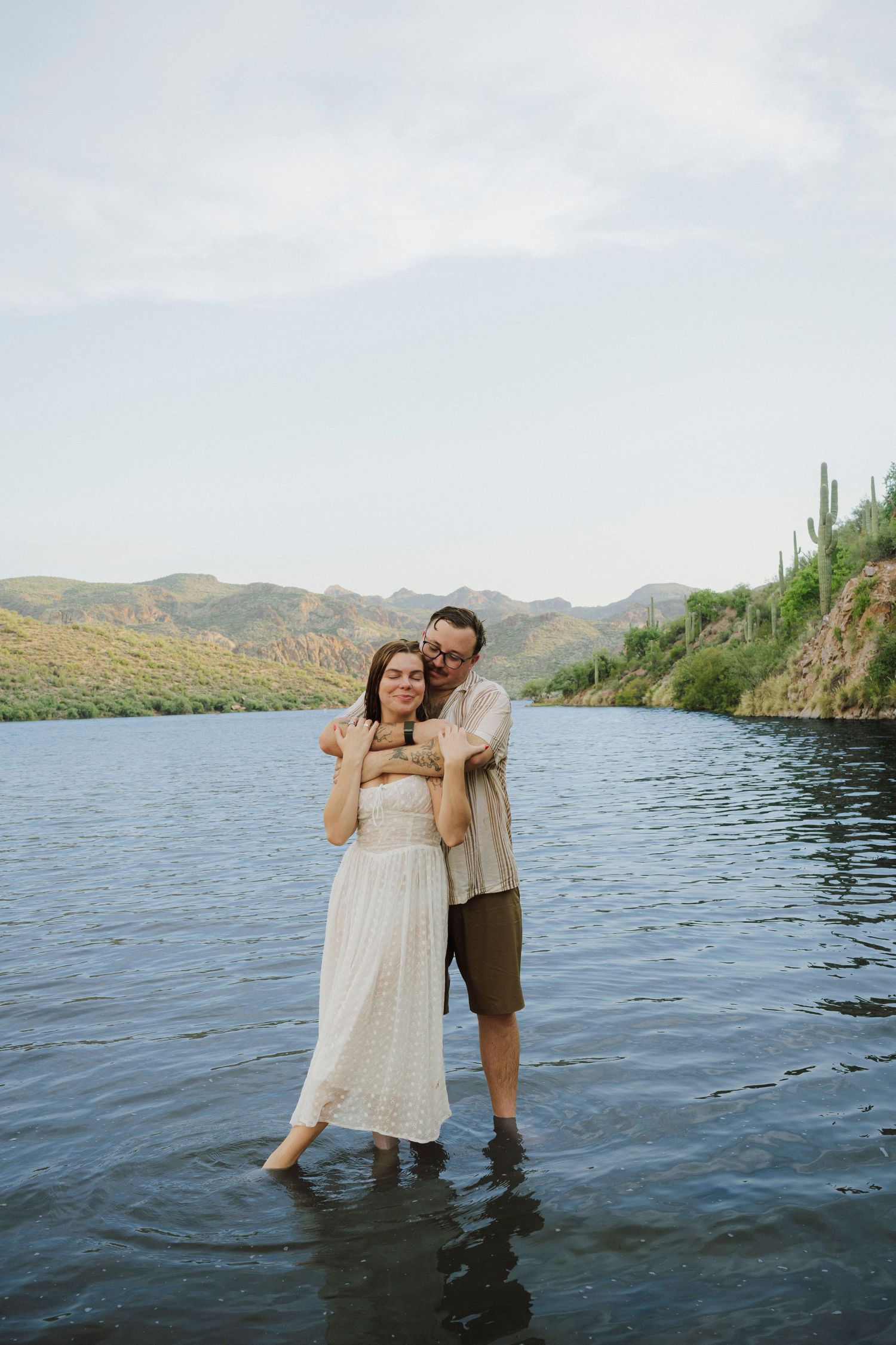 Couple standing in a lake embracing, wearing white dress and shorts, with green hills and cacti in background.