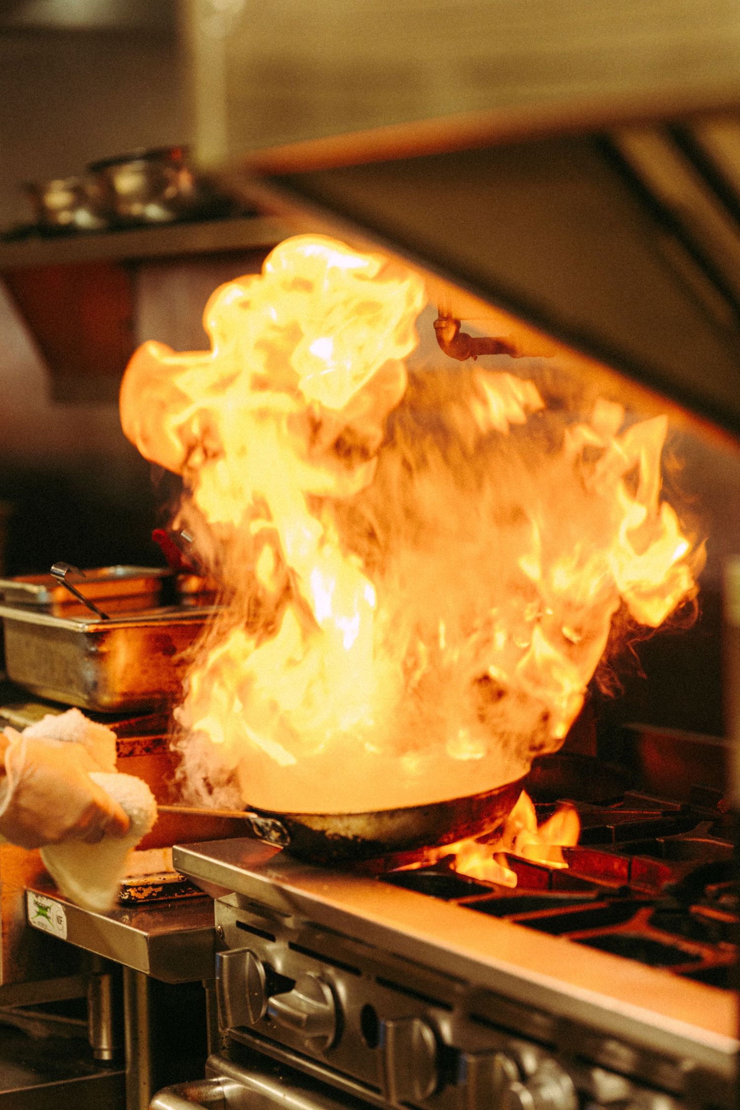 Chef flambéing food in a pan with dramatic flames on a commercial kitchen stove.