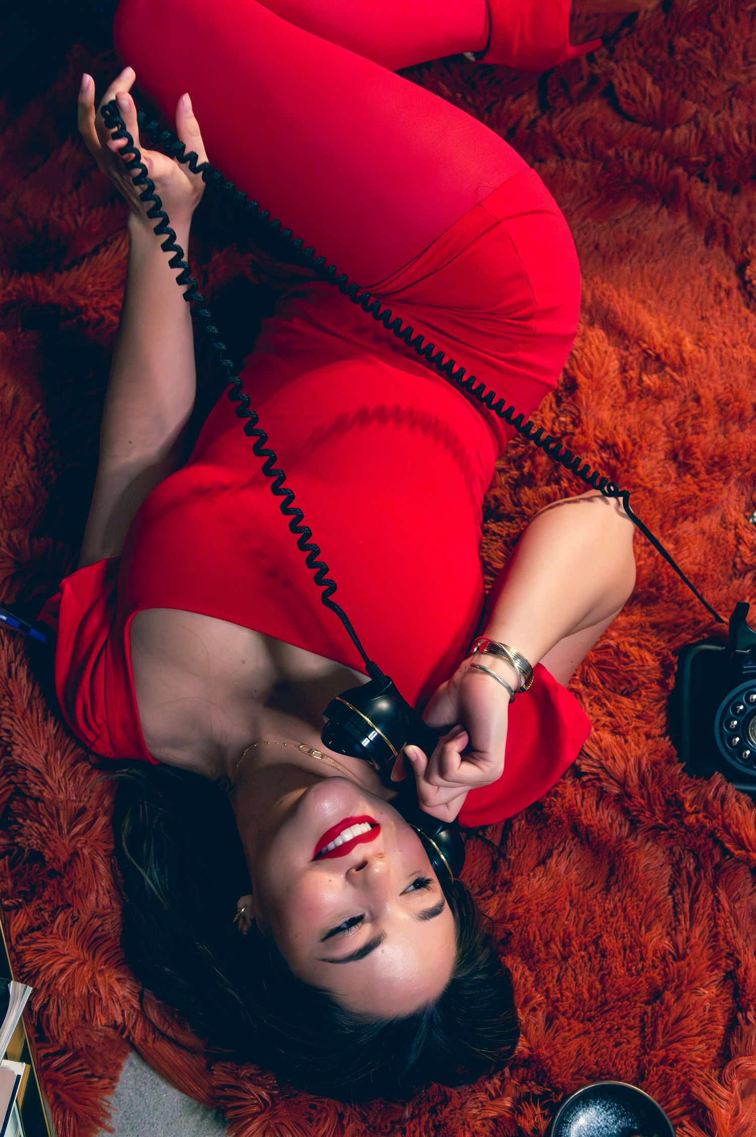 Woman in red outfit lying on orange fur rug talking on a vintage rotary telephone.