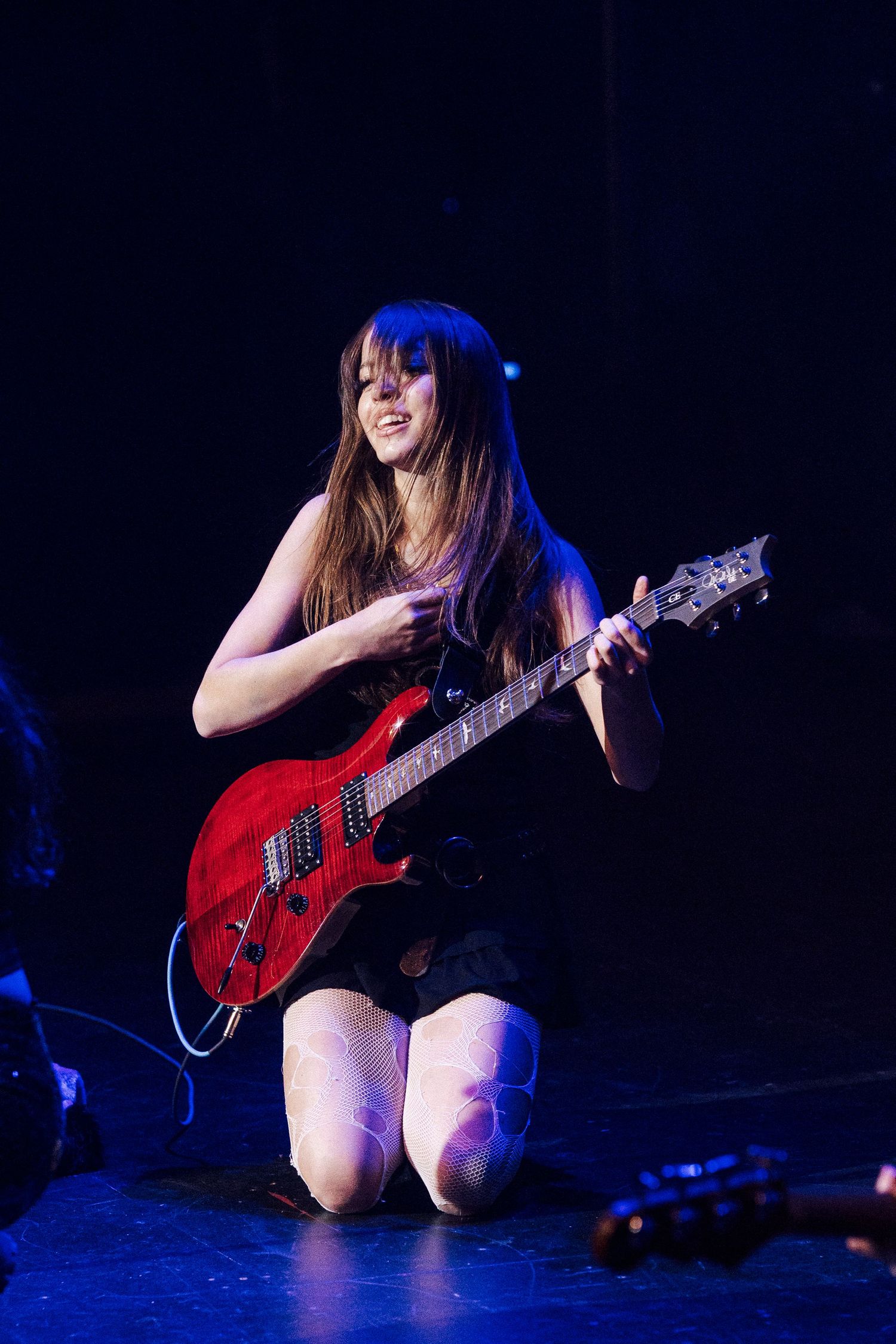 Female guitarist kneeling on stage playing a red electric guitar under blue stage lighting.